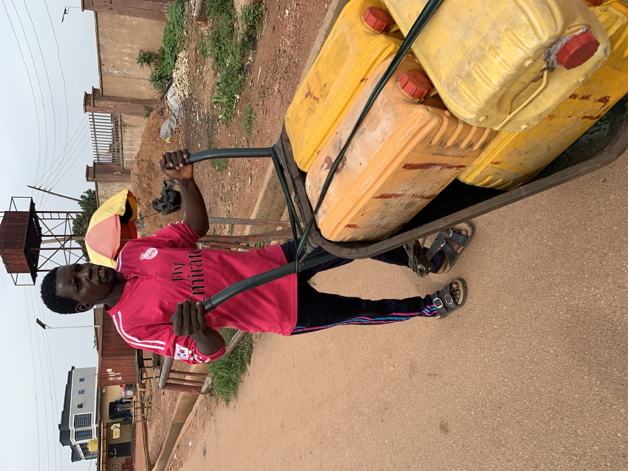 A man, with dark skin and a short haircut, is pushing a cart laden with yellow jerrycans filled with liquid. He is wearing a bright pink t-shirt with white stripes on the sleeves and black pants with colored stripes. He has on black sandals. The cart appears to be made of metal and has two green straps holding the jerrycans in place. The man is standing on a dirt road, and behind him, there are several buildings, a fence, and piles of dirt and debris. The sky is overcast. This scene is taking place in the city of Jos, Nigeria.