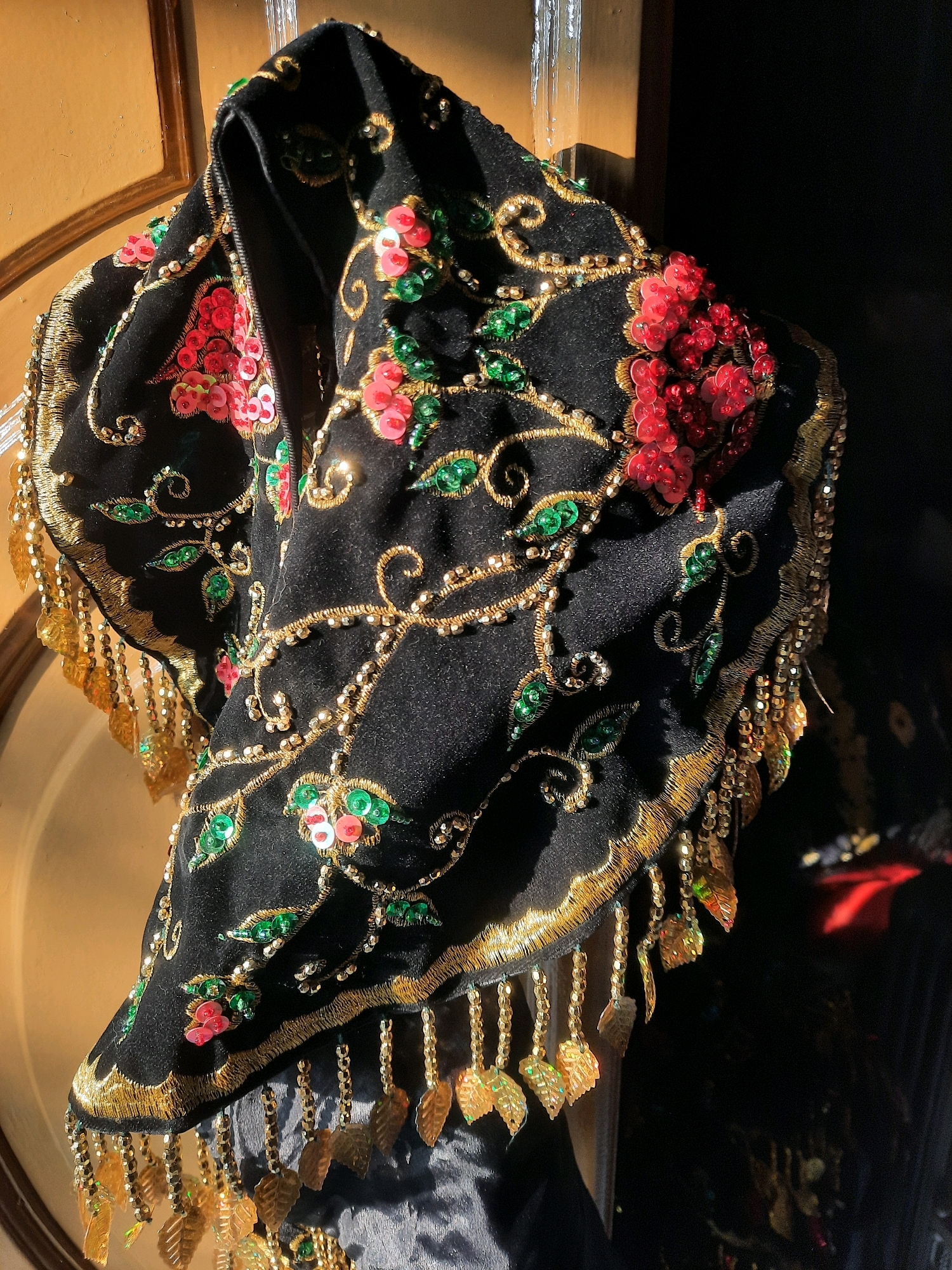 A black velvet scarf with intricate gold embroidery and colorful sequins is draped over a wooden surface. The scarf features floral motifs with red and pink sequins and green sequined leaves. Gold beads and chains are also incorporated into the design, and a fringe of gold beads and leaf-shaped ornaments hangs from the edges. The background is dark, with hints of a wooden frame and reflections. The overall impression is one of a richly decorated textile. This image does not provide any information about its location in East Jakarta, Indonesia.