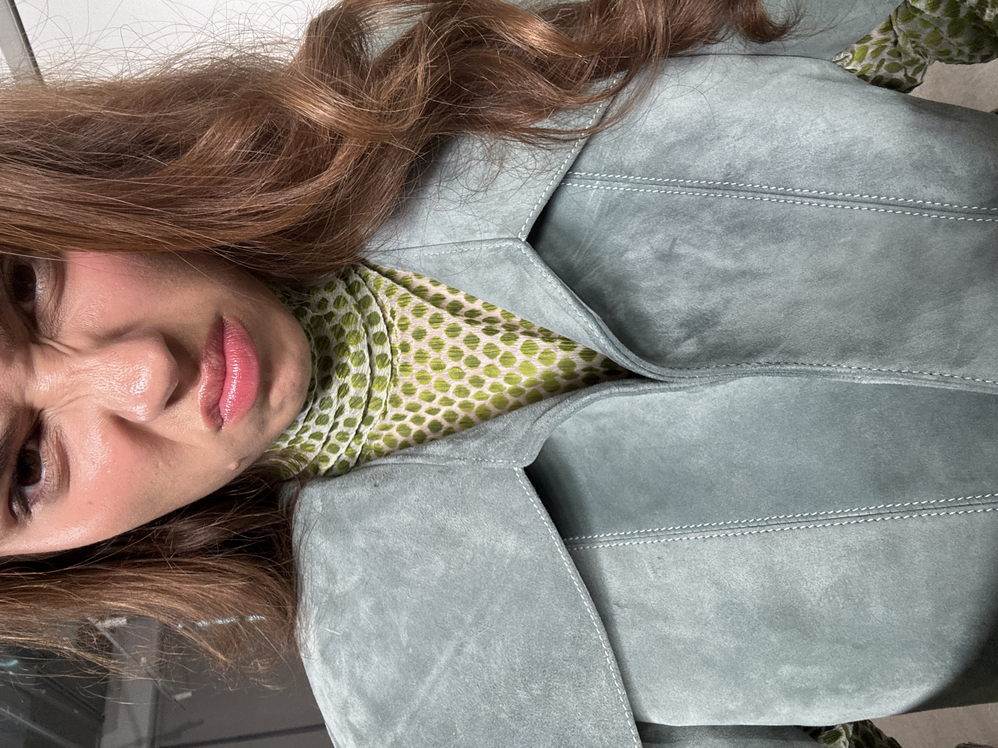 A close-up, over-the-shoulder shot focuses on the upper torso and face of a young woman wearing a pale blue suede jacket over a green and white patterned shirt. Her face is contorted into a disgusted or displeased expression, with her eyebrows furrowed and her lips pursed. Her long, wavy brown hair falls over her right shoulder. The background appears to be an indoor setting, possibly a studio or backstage area, with a plain white wall visible. The scene does not appear to be located in Rishon LeZion, Israel; there are no visual cues suggesting this location.