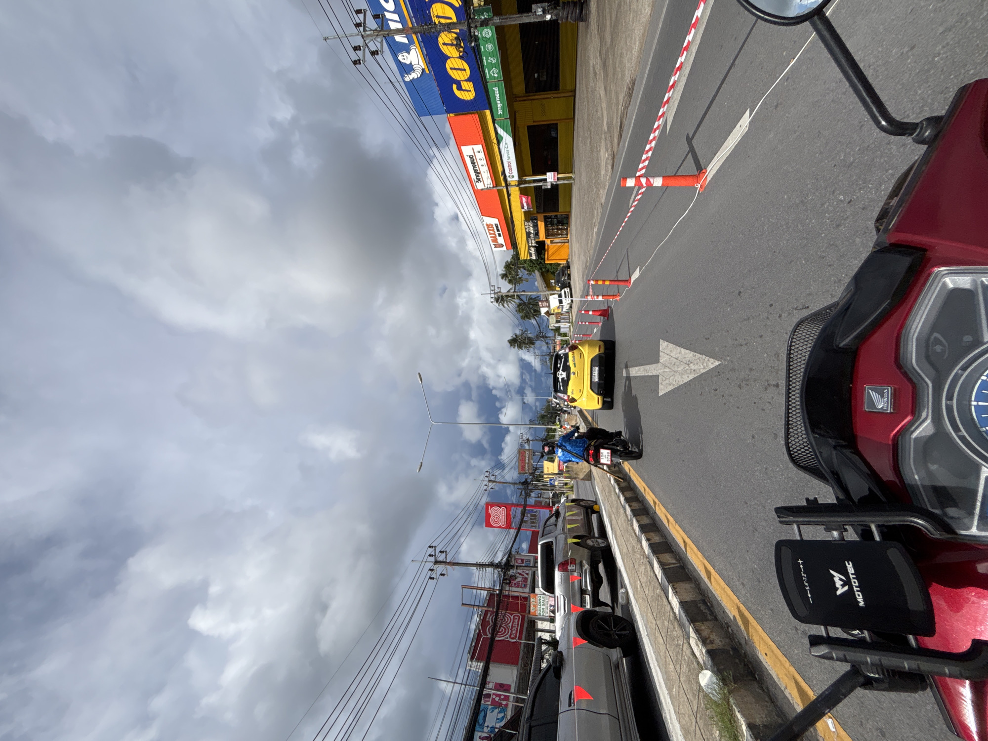 The image shows a view from the perspective of a motorcyclist on a road in Rawai, Thailand. The sky is heavily overcast with dark, dramatic clouds, suggesting an approaching storm. On the right side of the road, there are several commercial buildings with large signage, including "MAXIS TIRE SERVICE" and "GOODYEAR." There are traffic cones and a red and white barrier tape along the edge of the road, indicating some sort of roadwork or lane closure. Several vehicles are visible on the road, including a silver pickup truck, a silver car, a yellow car, and a motorcycle with a rider wearing a blue jacket and helmet. The road surface is asphalt, with clear lane markings and a yellow line demarcating the edge of the traffic lane. The overall scene depicts a busy street with ongoing traffic under a moody, cloudy sky.