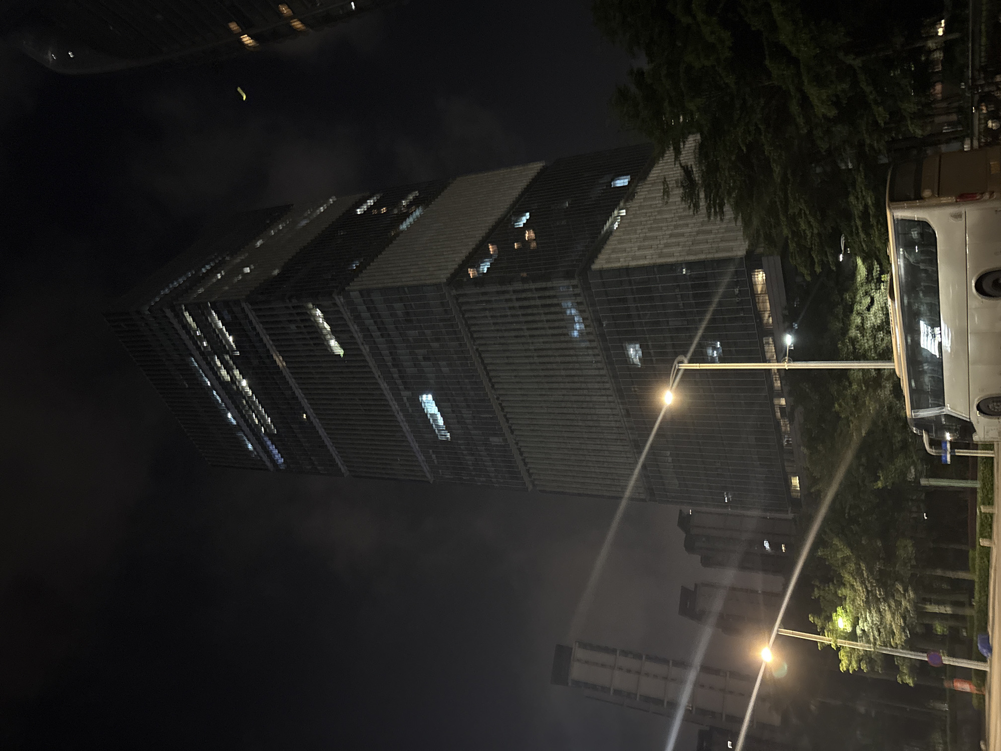 The image depicts a nighttime scene in a city, likely Haizhu District, China. A tall, modern skyscraper with a unique architectural design dominates the frame. The building features a stepped, segmented facade with alternating dark and light panels. Many windows are illuminated, suggesting occupancy and activity within the structure.
In the foreground and to the right, a white vehicle, possibly a van or RV, is parked. Next to it, lush green trees provide a natural contrast to the urban environment. Streetlights cast a warm glow on the scene, illuminating the pavement and creating streaks of light across the image due to the camera's perspective and movement. In the background, other buildings and city lights are visible, indicating a dense urban setting. The sky is dark, suggesting it is nighttime, and some cloud cover is present, obscuring the stars. The overall atmosphere is one of a bustling, illuminated city at night, with a focus on the striking architecture of the skyscraper. No people are clearly discernible in the image.
