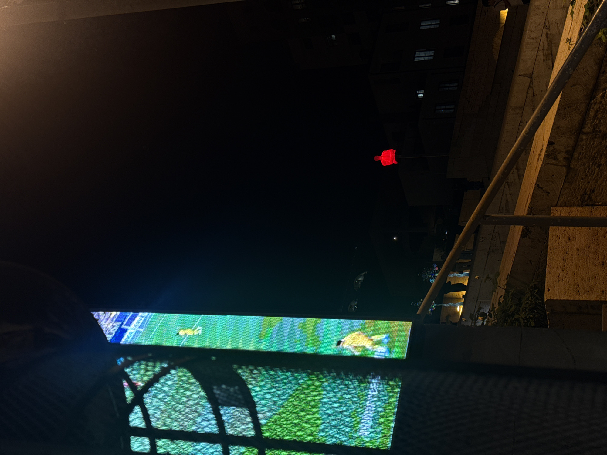 The image depicts a night scene, seemingly in Yerevan, Armenia. A television screen is visible, displaying a football match. The screen is positioned in what appears to be an outdoor setting, potentially a cafe or balcony. In the distance, buildings are silhouetted against the dark sky, punctuated by a red light atop one structure. A structure with a railing is present on the right side. The image is taken from a low angle, looking up towards the screen and the cityscape.