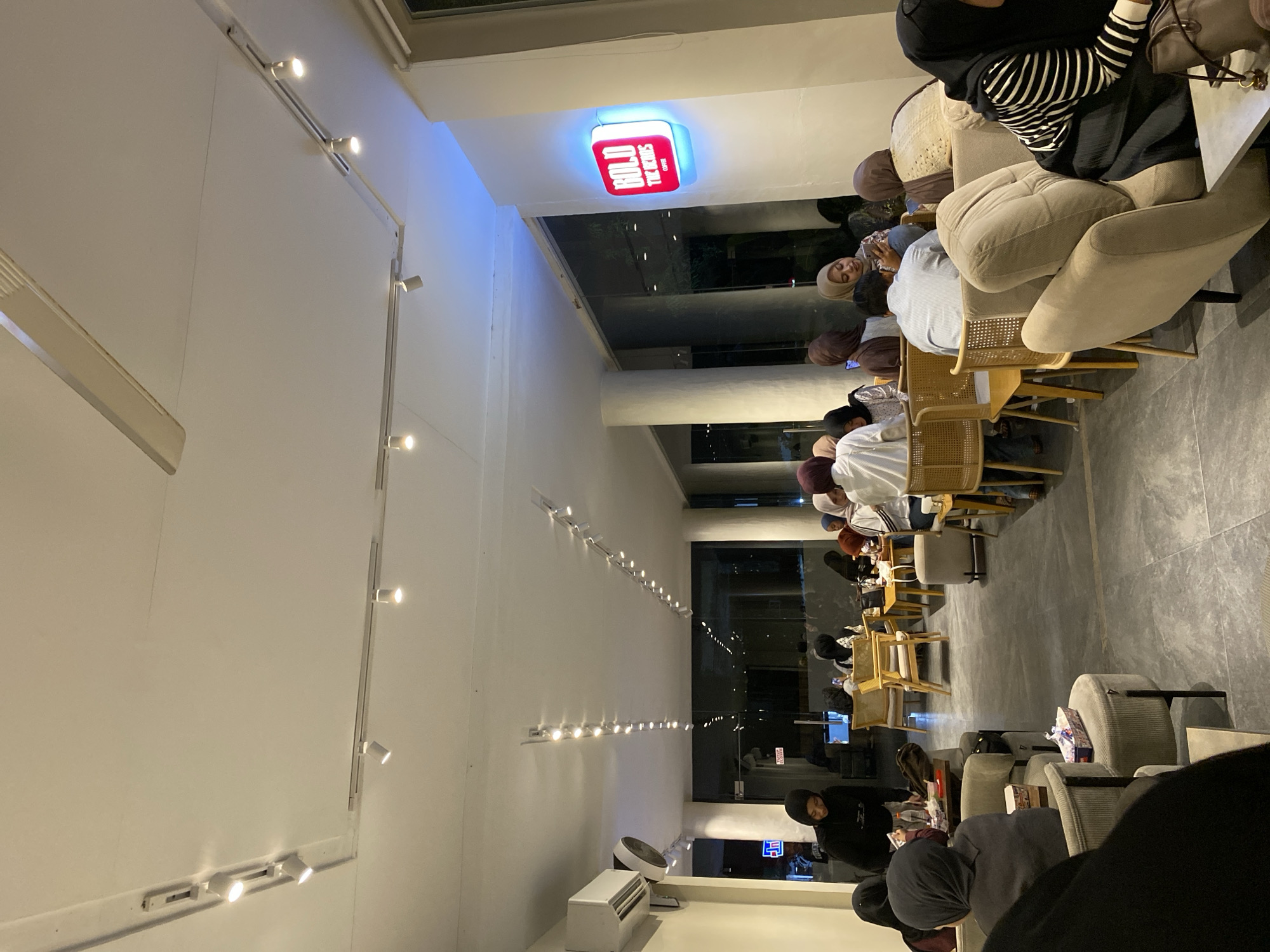 The image shows the interior of the Bold The Kings Coffee shop in Lhokseumawe, Indonesia. The coffee shop has a long, narrow layout with seating on both sides. People are seated at tables, mostly in groups, some wearing hijabs. The shop is lit by recessed lighting on the ceiling and track lighting along the walls. An air conditioning unit is visible near the back of the shop. A sign with the coffee shop's name is located near the front. The scene appears to be during the evening.