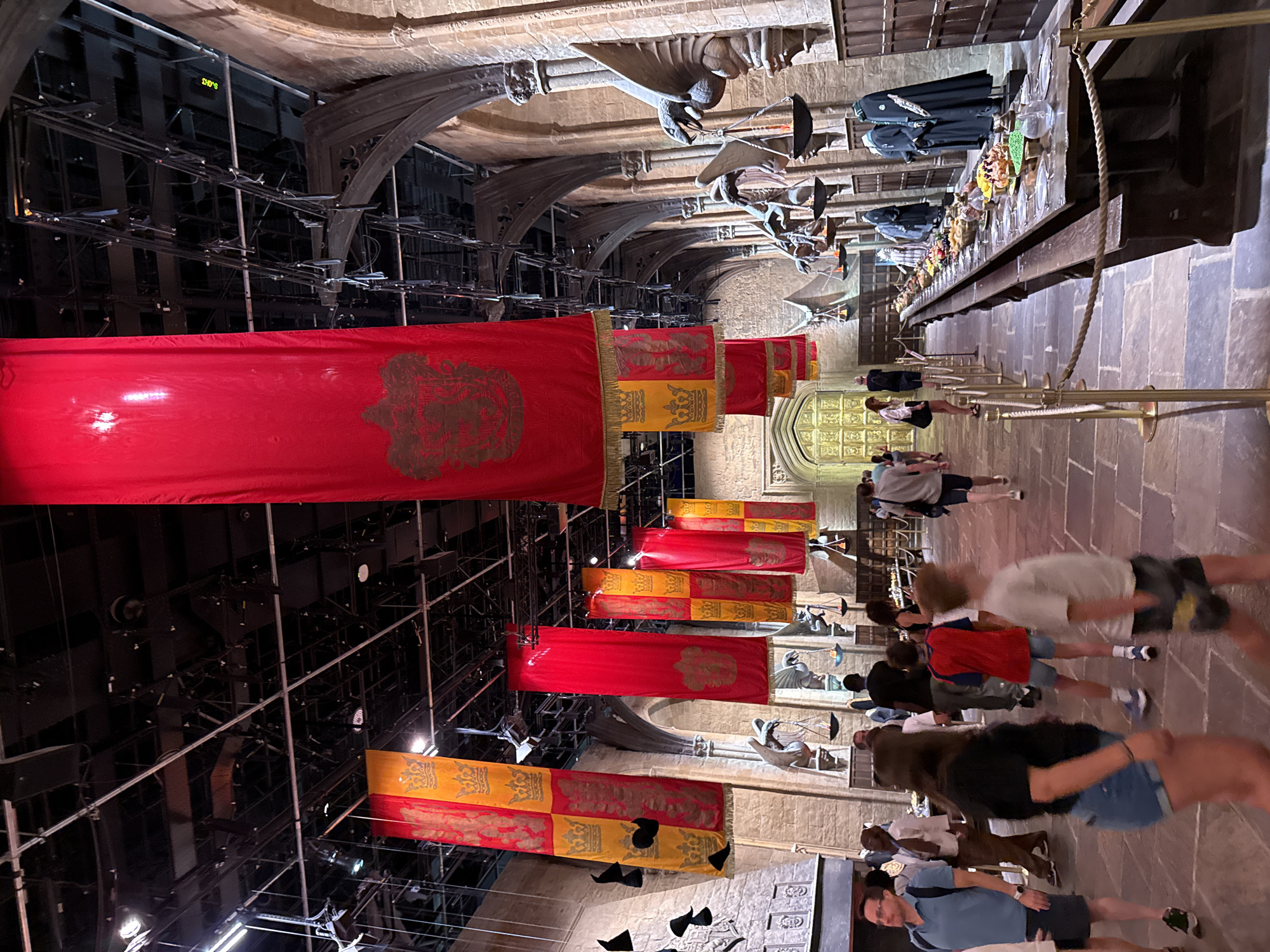 This is an indoor scene at Warner Bros. Studio Tour London – The Making of Harry Potter in Watford, United Kingdom. The location appears to be the Great Hall, decorated with house banners and a long table laden with food. The banners, in red and yellow, feature crests with crowns and lions. Several people are walking through the hall, some are looking at the table, and others are walking in different directions. The hall has stone walls, arched doorways, and a high ceiling with exposed metalwork and lighting rigs. Gargoyle statues are mounted on the walls, holding braziers with flames. The lighting is somewhat dim, creating an atmospheric effect.