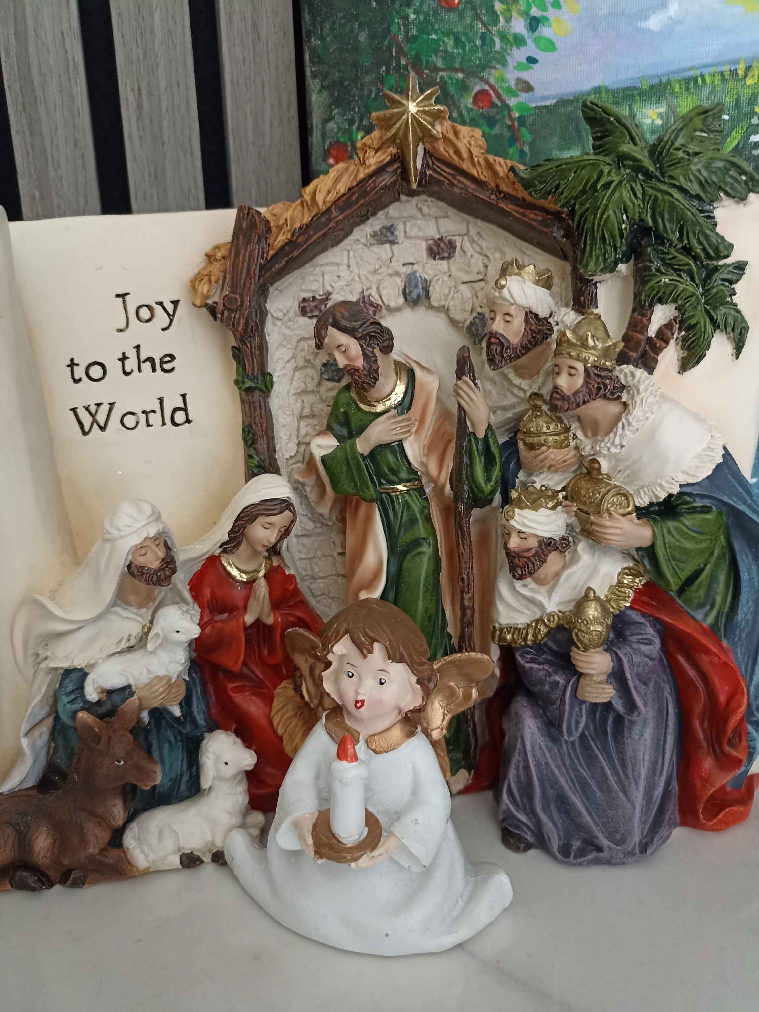 This is a close-up, eye-level, indoor shot of a nativity scene figurine. The figurine depicts a stable with a thatched roof and stone walls. Inside the stable, a man with a beard and a green robe, likely Joseph, stands with a wooden staff. To his right, a woman with a red robe and a white head covering, likely Mary, kneels with her hands clasped in prayer. Beside her, a man with a white head covering and a blue robe, also bearded, holds a lamb. To the right of Joseph, three figures, presumably the Three Wise Men, are present. They wear crowns and are dressed in robes of various colors, including purple, blue, and white. Each of them holds a golden vessel. In front of the stable, an angel figurine with golden wings and a white robe holds a lit candle. A brown donkey is positioned to the left of the stable, and another white lamb is in front of Mary. A scroll with the words "Joy to the World" is visible to the left of the stable. The background consists of a painted landscape with trees and a sky. The figurine is placed on a white surface. There is no indication of the location being in Doirí Beaga, Ireland.