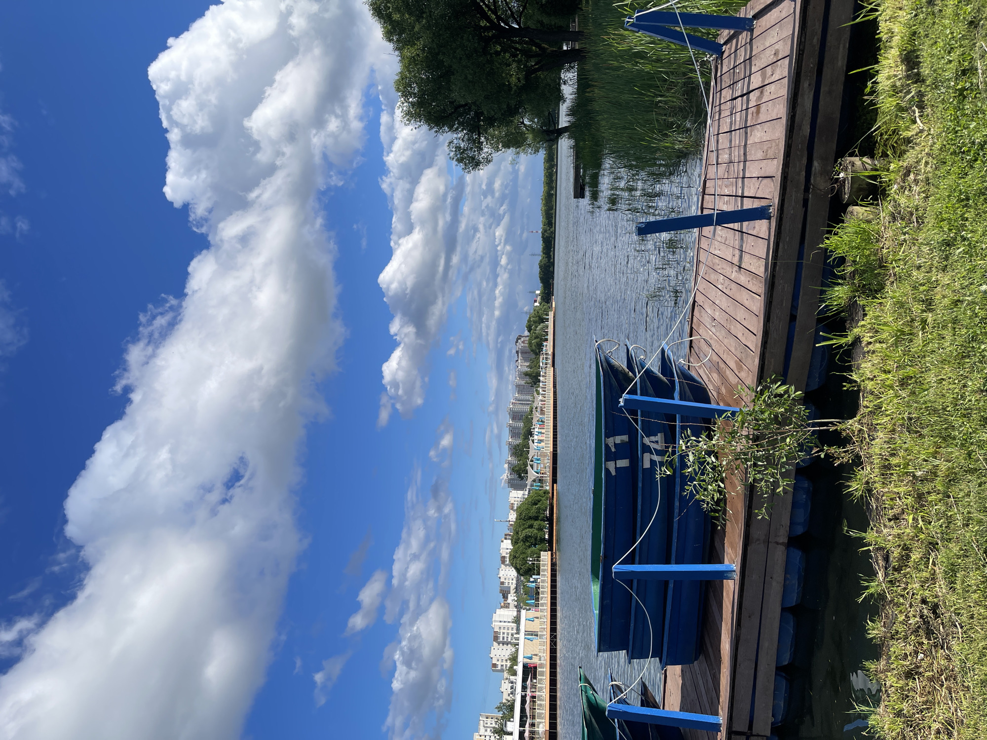 This image depicts a serene lakeside scene in Moscow, Russia, specifically at the Izmailovo Kremlin or the area surrounding it. In the foreground, a wooden pier juts out into the calm water, laden with stacked blue rowboats, some marked with numbers "11" and "74." The pier is secured by blue posts and ropes, and the water reflects the clear blue sky dotted with fluffy white clouds. Lush green grass lines the edge of the pier, with a small sapling growing near the water.
Across the lake, a cityscape of modern, white buildings is visible, interspersed with trees. A boardwalk or promenade runs along the waterfront, with awnings and structures suggesting a recreational area. The background features a dense line of trees separating the water from the urban landscape, and in the distance, what appears to be a red and white pylon or tower can be seen against the horizon. The overall atmosphere is one of tranquility and leisure on a sunny day.