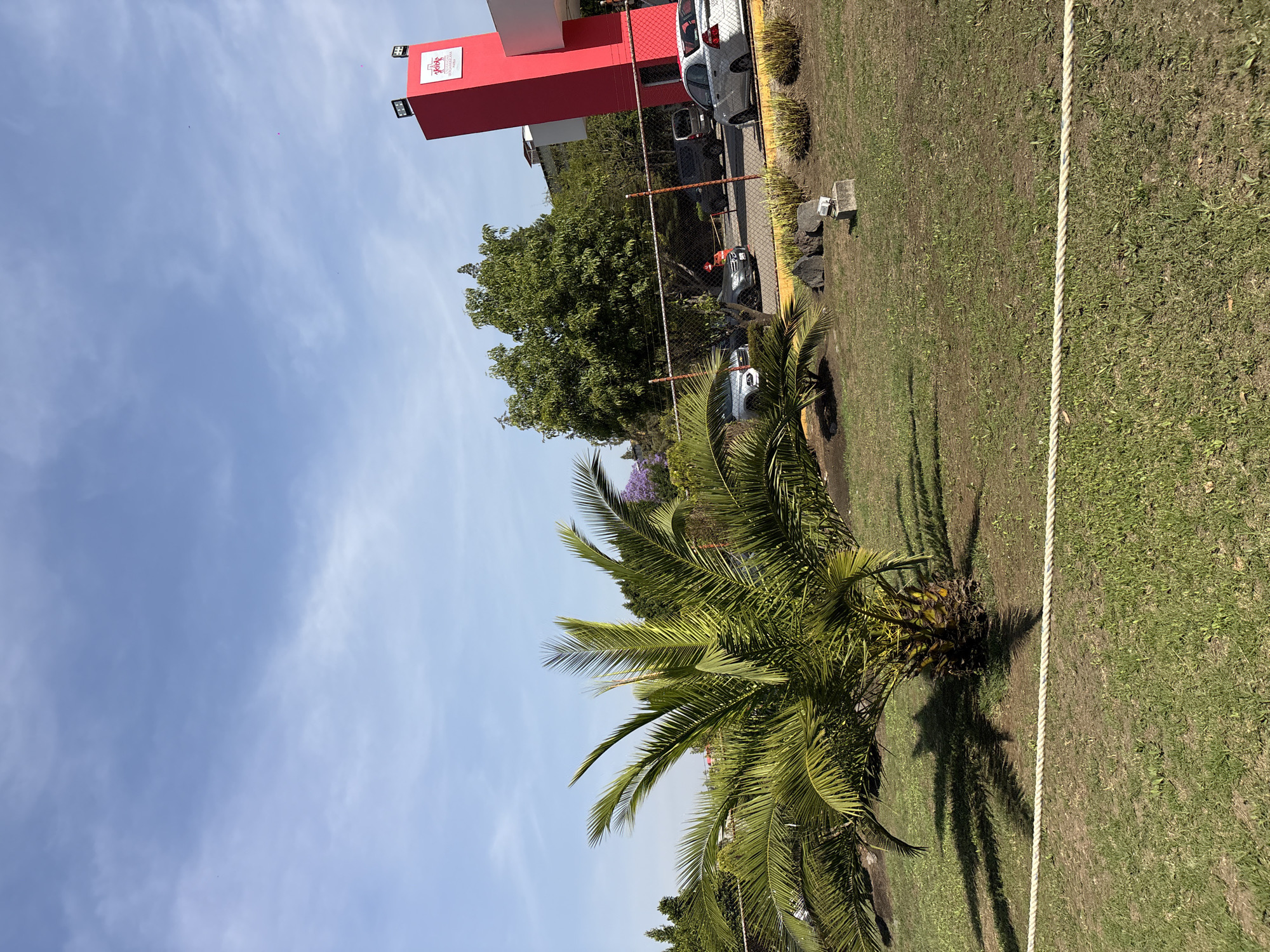 This image, taken in San Bernardino Tlaxcalancingo, Mexico, presents an outdoor scene under bright, sunny conditions, likely late morning or early afternoon given the strong shadows. The sky is a clear blue with scattered, wispy white clouds.
In the immediate foreground, a vibrant green grass field dominates the lower half of the frame. A single, taut white rope runs across the grass, appearing to mark a boundary or demarcation line. To the left of this rope, a mature palm tree with luxuriant green fronds stands prominently, casting a distinct shadow on the grass.
The midground features a paved area, likely a parking lot or driveway, where several vehicles are parked, including a white car and a dark-colored SUV or truck. A chain-link fence separates the grassy expanse from this paved section and a dense cluster of other trees. Among these trees, hints of purple blossoms suggest the presence of a Jacaranda.
In the background, a striking red building with a modern, blocky design is visible. Attached to its side is a white sign featuring a red logo and text. The legible text on the sign reads "LA CONTRALORÍA" at the top and "UNIVERSITARIA" below, indicating it is likely a university's comptroller's office. Overhead lights are mounted on the building.
The scene is devoid of people or discernible activity, conveying a calm and static atmosphere typical of a well-maintained institutional or commercial campus.