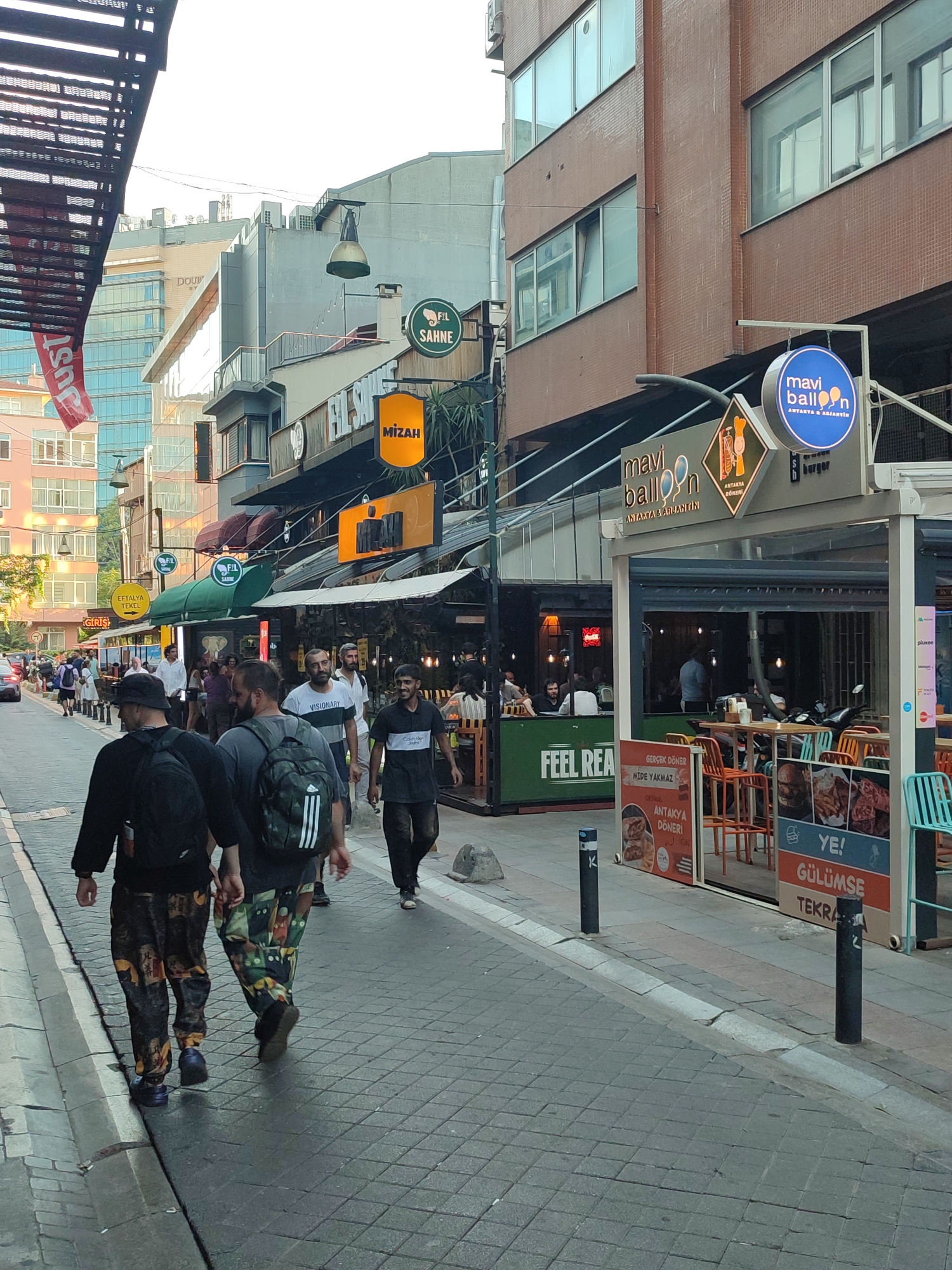 The image captures a street scene in Kadıköy, Turkey, likely during daylight. The foreground shows the backs of two men walking away from the camera on a paved sidewalk. Both are wearing patterned trousers and backpacks. The man on the left has a black backpack and dark clothing, while the man on the right has a camouflage backpack and a grey shirt. To the right of the sidewalk, a restaurant or cafe area is visible with outdoor seating, tables, and chairs. Several signs are displayed, including one for "Mavi Balloon" and another with images of food. In the background, buildings line the street, featuring a mix of architectural styles. Signs for businesses like "Fil Sahne" and "Mizah" are also visible. People are seen walking and dining in the area, suggesting a bustling urban environment. The overall impression is of a lively commercial street in a Turkish city.