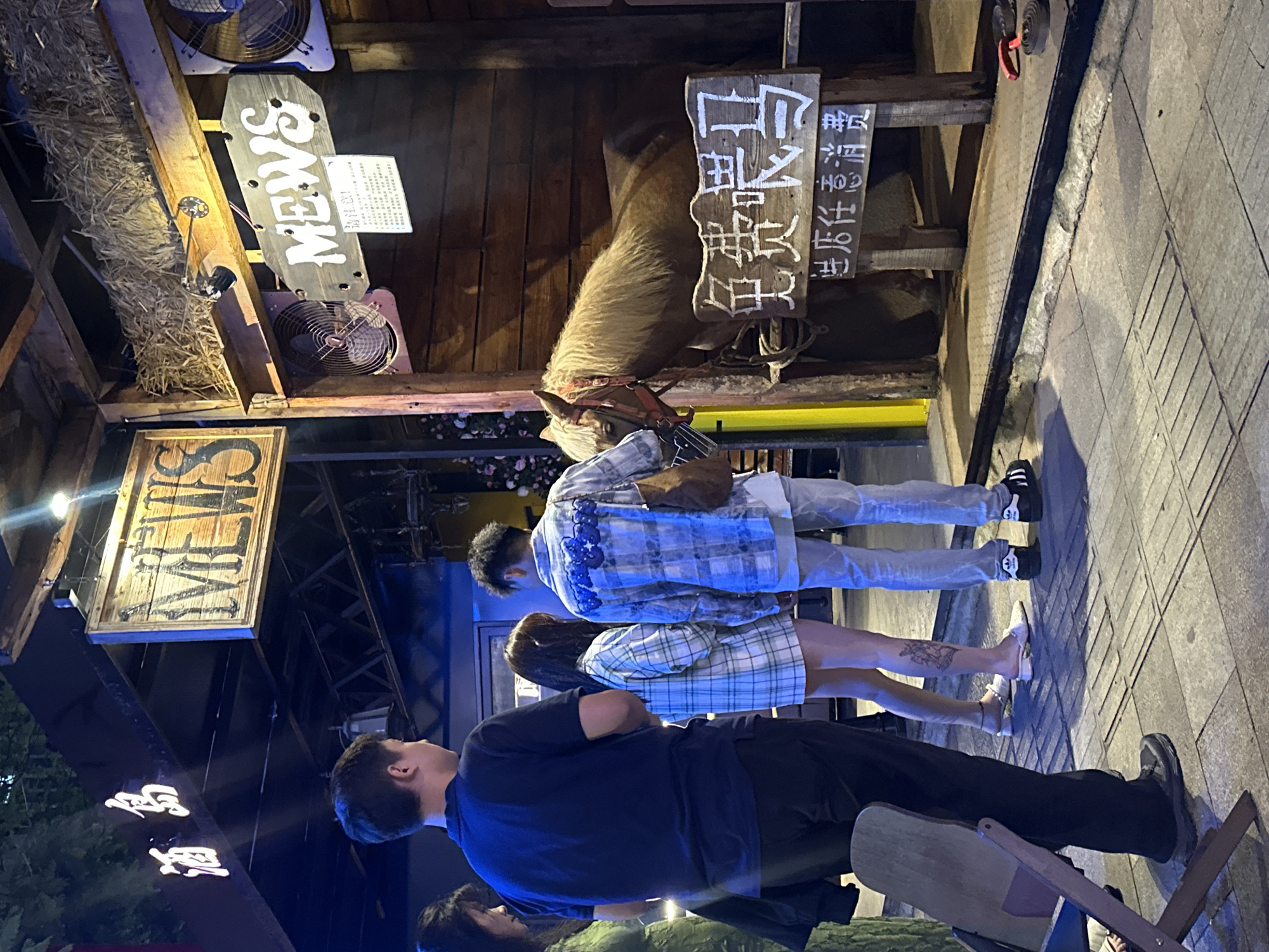 In the city of Chongqing High-tech Zone, China, at night, several individuals stand before a wooden enclosure containing a light brown horse. The enclosure has a rustic appearance, with straw visible at the top and wooden planks forming the walls. Two signs are prominently displayed: one above the enclosure reads "MEWS" in a stylized font, and another, closer to the horse, features Chinese characters that translate to "Free Horse Riding" and "Any consumption in the store." The individuals are observing the horse, with one person extending their hand towards it, possibly offering food. The ground is paved with tiles. The lighting is dim, casting shadows and highlighting the subjects.