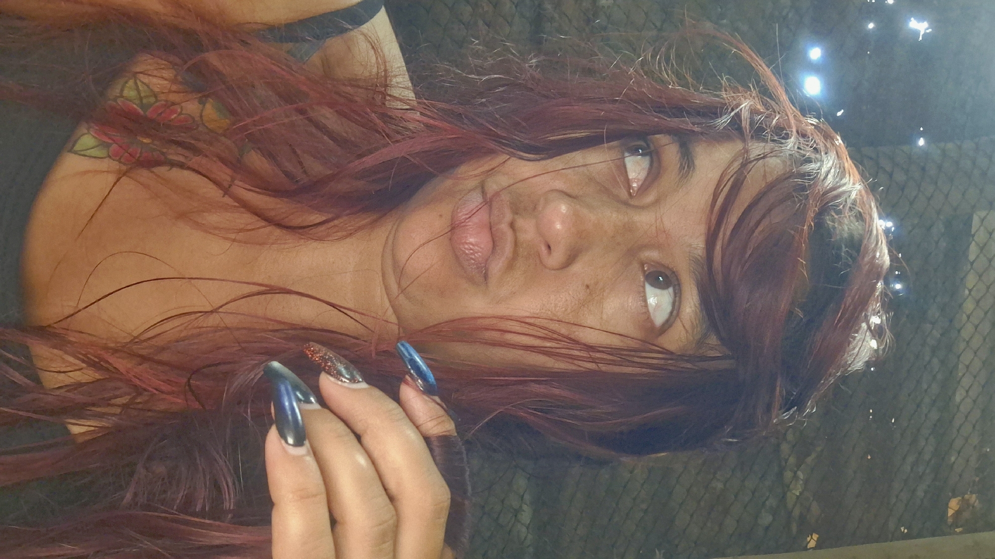 In Vallecito, Venezuela, a woman with auburn hair and a tattoo on her chest is captured in a selfie. She is looking to her left with pursed lips and her hand is raised, showing off her long, dark blue nails with glitter accents. The background appears to be a chain-link fence with some blurred lights, suggesting an outdoor or semi-outdoor setting at night. The overall lighting is dim and atmospheric.