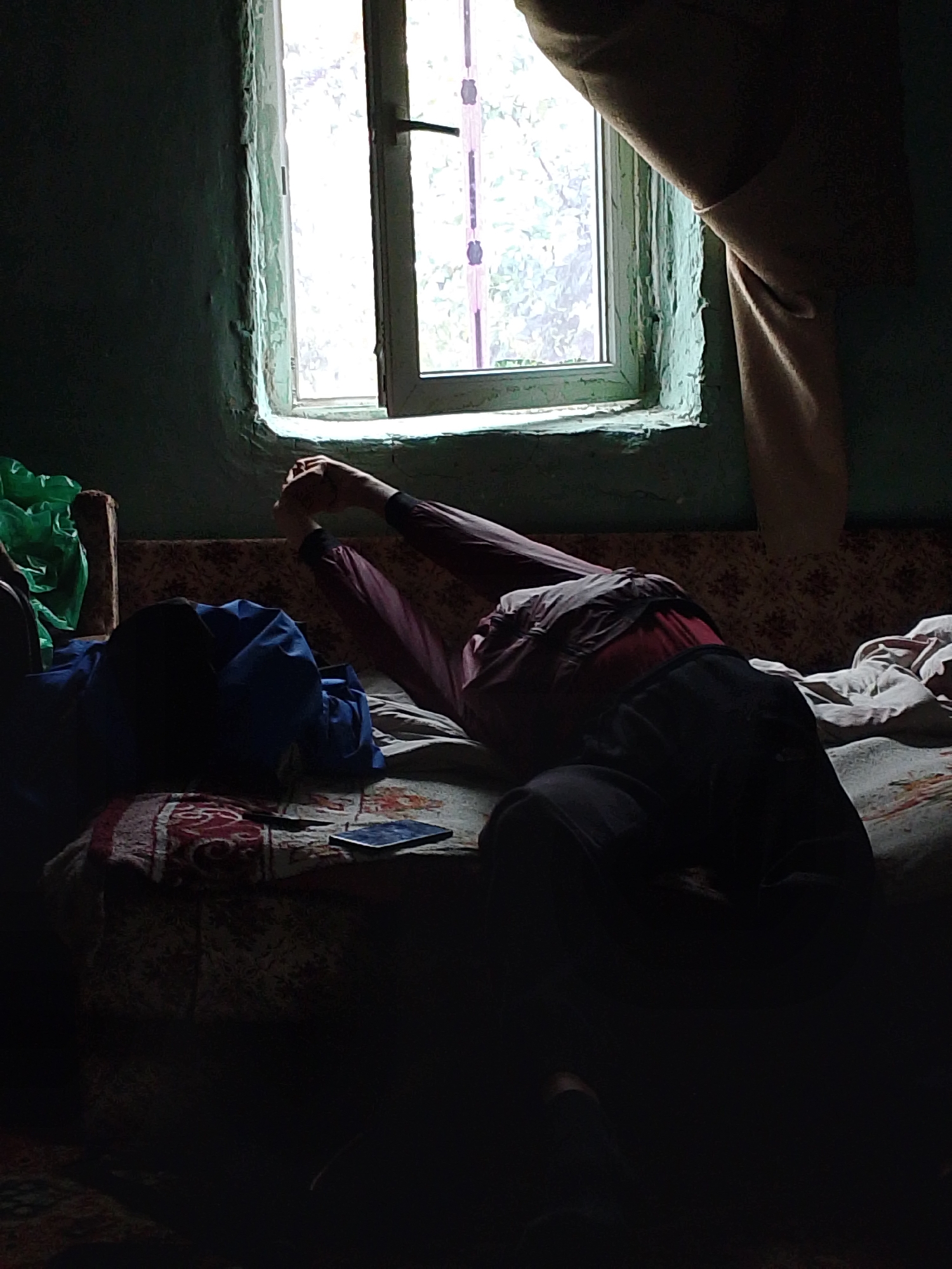 A person is lying on a couch, positioned under a window. The individual is wearing a long-sleeved shirt and dark pants. Their hands are raised above their head. The room is dimly lit with natural light coming through the window. A curtain hangs to the side of the window. A blue bag is on the couch near the person. A mobile phone rests on the couch. The interior walls appear to be peeling and slightly damaged. The location is Ustyvytsia, Ukraine.