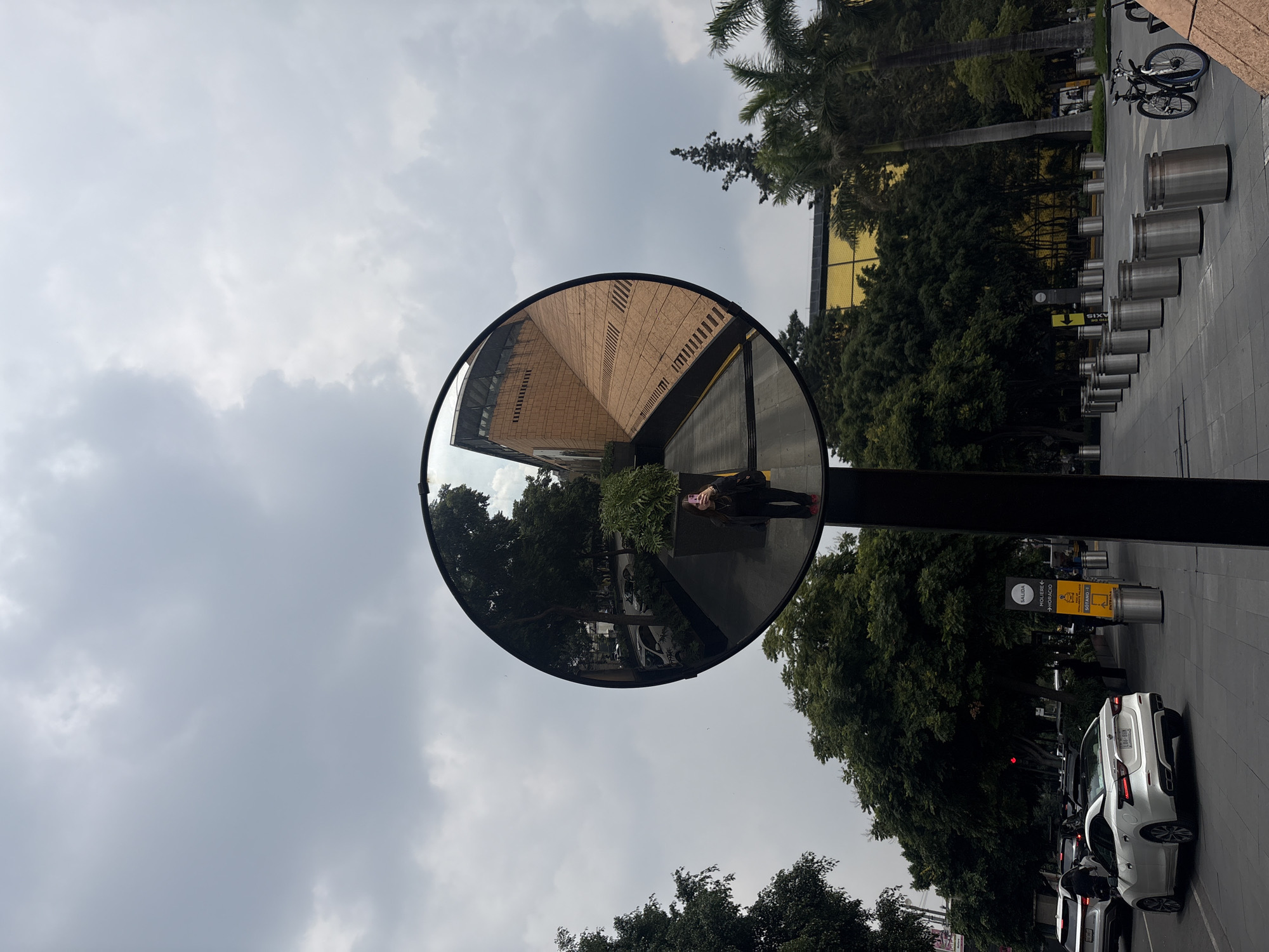 The image shows a convex security mirror on a city street in Mexico City, Mexico. The mirror reflects a person taking a selfie, the surrounding trees, buildings, and a street with cars. In the foreground, there is a sidewalk with metal trash receptacles and bicycles. A white car is parked on the street, and other cars are visible in the background. The sky is cloudy. The architecture of the buildings suggests an urban setting, with a modern building featuring a brick facade and a building with a yellow reflective surface. The overall scene depicts an everyday urban environment in Mexico City, captured through the reflective surface of a security mirror.