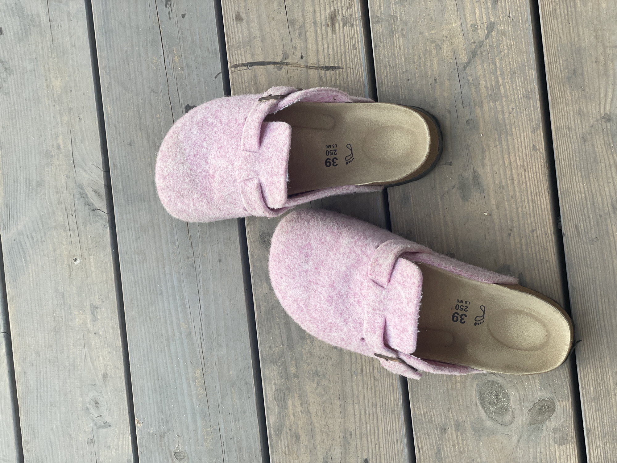 A pair of pink fuzzy clogs are displayed on a wooden deck. The clogs have a strap with a buckle across the top. The insoles of the clogs are visible, showing markings for size 39. The wooden deck planks are weathered and gray.