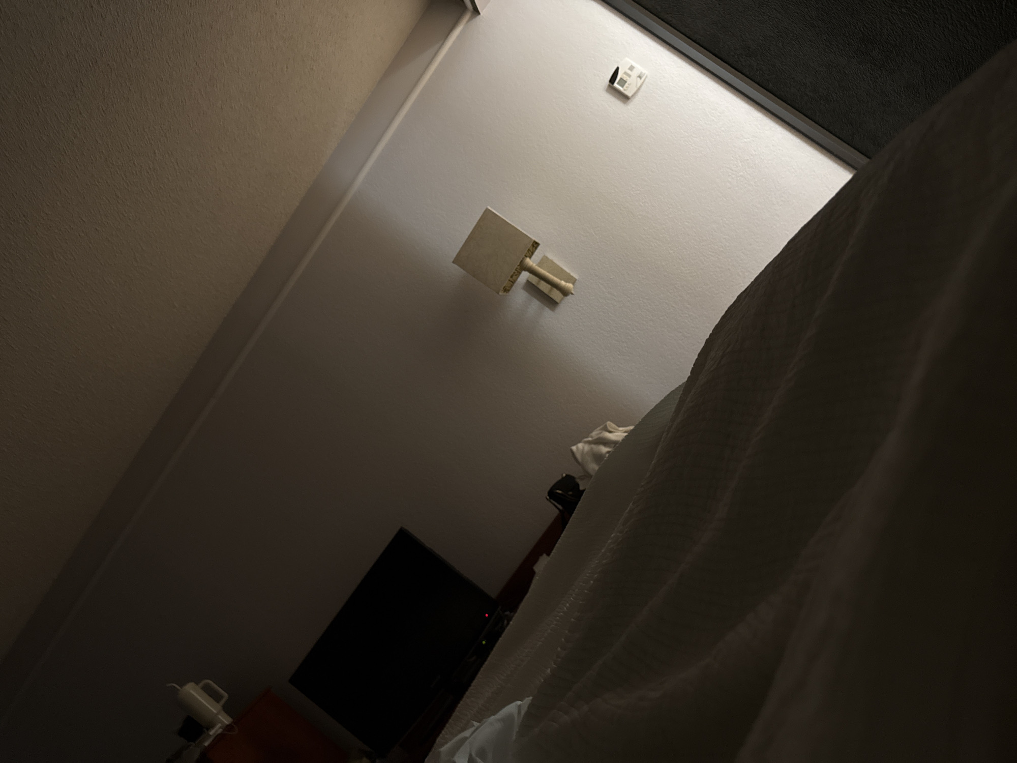 This image depicts a dimly lit interior scene, likely a hotel room, in San Antonio, United States of America. The scene is dominated by a bed covered in white sheets, filling the right side of the frame. On the wall, a modern, square wall lamp is visible, and a thermostat control panel is located near the top right corner. A television is visible on a stand against the wall. The overall ambiance is subdued, suggesting nighttime or low ambient light.