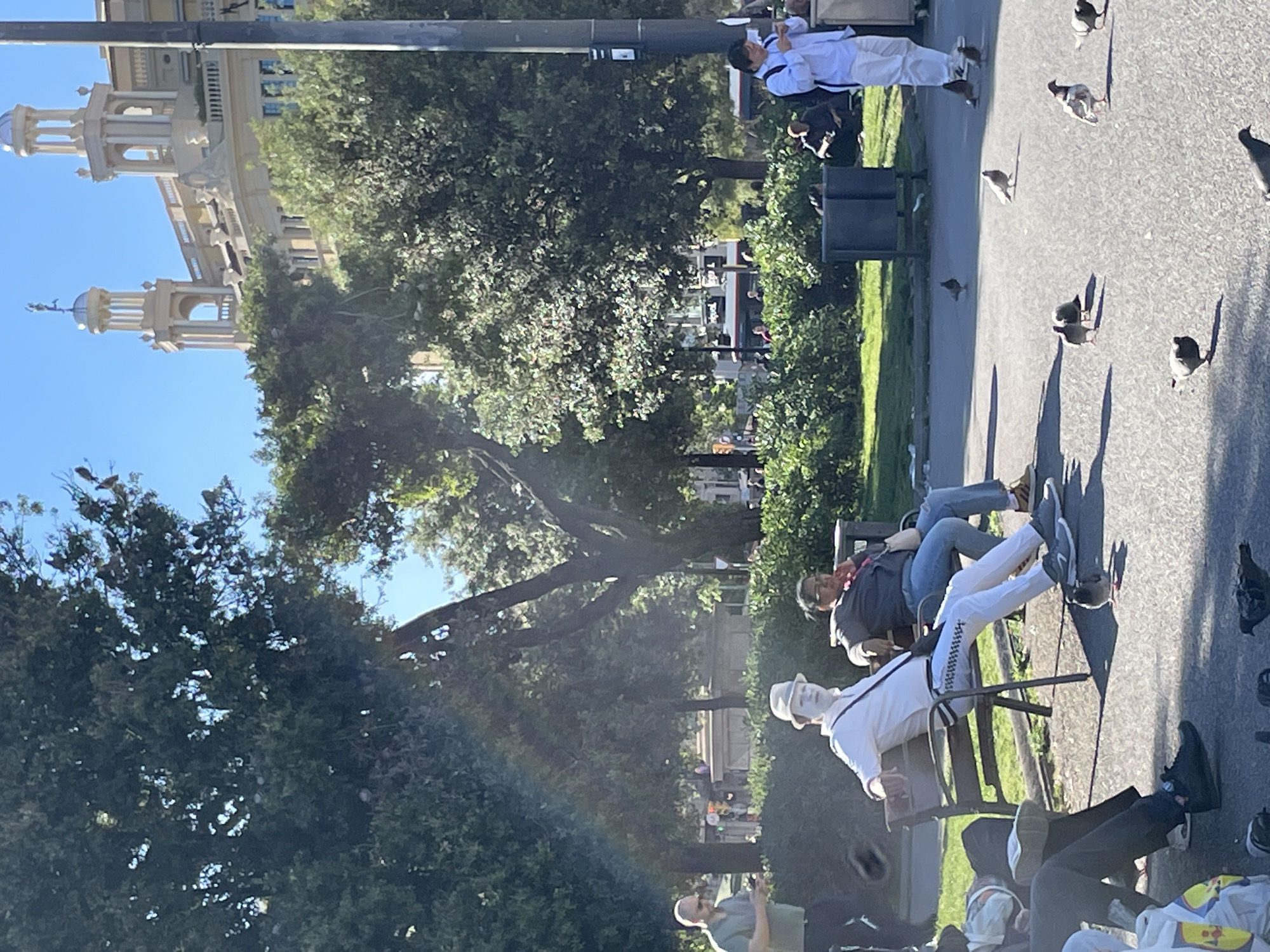 The image captures a scene in Barcelona, Spain. A group of people is sitting on benches in a park or plaza. In the background, a building with towers is visible. Some individuals are lying on the benches, while others are seated. Pigeons are scattered on the pavement. The lighting suggests it is daytime. The scene appears to be a public space where people are relaxing.