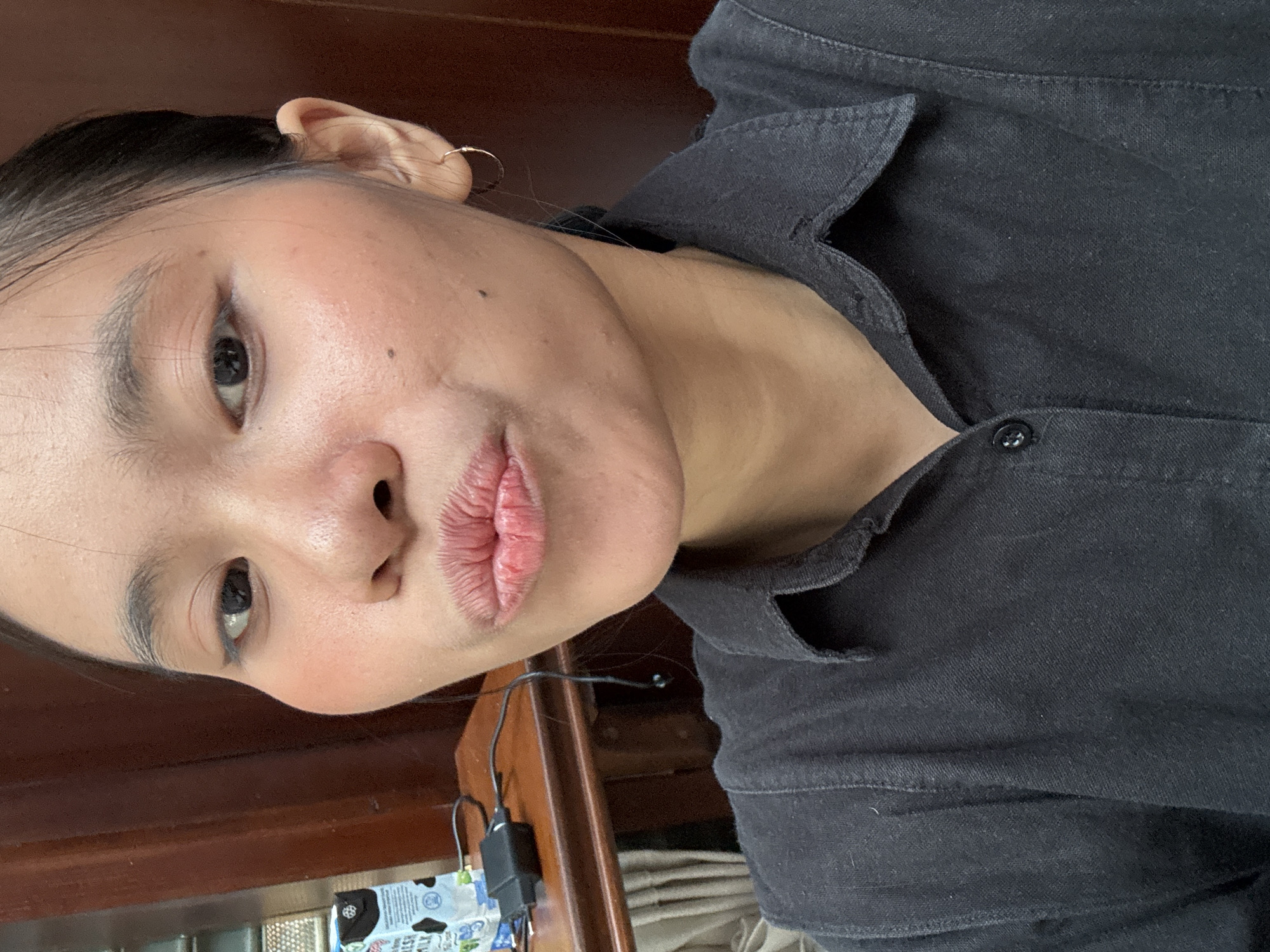 A close-up selfie shows a young woman with dark hair pulled back, puckering her lips. She wears a dark collared shirt. Her right ear has a small hoop earring. Behind her, a wooden desk holds a carton of milk and a black charging adapter with wires. The background is a dark brown, wooden surface, likely a wall or furniture. The lighting is bright and direct, casting subtle shadows. The scene appears to be indoors, likely in a residential setting within the Kebayoran Baru subdistrict of South Jakarta, Indonesia, suggested by the style of furniture and the commonality of such personal items.