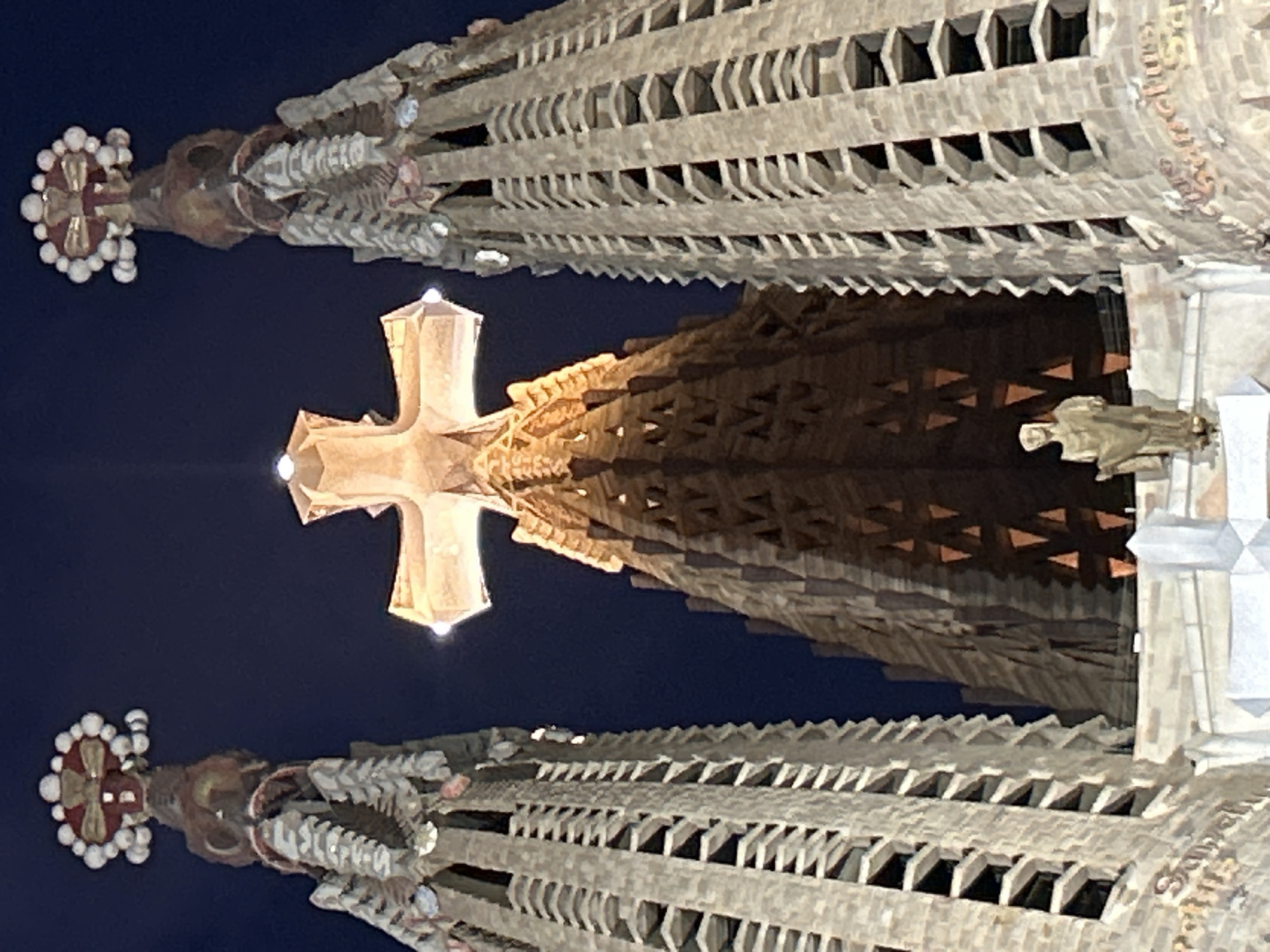 The image shows a low-angle, exterior shot of the Sagrada Familia basilica in Barcelona, Spain, at night. The sky is dark blue. The basilica's facade features three spires, with a large, illuminated cross prominently displayed between two of them. The spires are intricately detailed with stone carvings and are topped with decorative elements. The central spire, behind the cross, is also visible and appears to be conical in shape with illuminated triangular openings. In the lower part of the image, a statue of a robed figure is visible on a ledge. The architectural style is Gothic Revival, characterized by its complex geometric patterns and organic forms. The lighting emphasizes the grandeur and intricate details of the basilica.