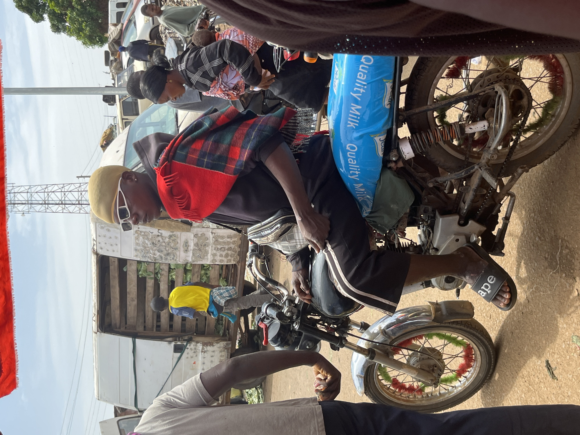 In this image, a group of people are gathered around a motorcycle in what appears to be an outdoor marketplace or a roadside area. The primary subject is a person sitting on the motorcycle, wearing a yellow cap, sunglasses, and a red and green plaid scarf draped around their shoulders. They are looking towards the left of the frame. Behind them, another person is partially visible, wearing a checkered shirt.
The motorcycle is black with chrome accents and has a large blue plastic bag attached to the back, on which the text "Quality Milk" is printed multiple times. The rear wheel of the motorcycle is decorated with red and green tinsel.
In the background, a white truck is visible, loaded with what looks like produce. The setting suggests a busy, possibly rural or semi-urban, environment, with hints of trees and sky in the upper portion of the image. The lighting indicates it is daytime. There is no strong indication of the weather conditions. The overall mood seems to be one of everyday activity and commerce. The image is oriented vertically, with the top of the scene appearing on the left.