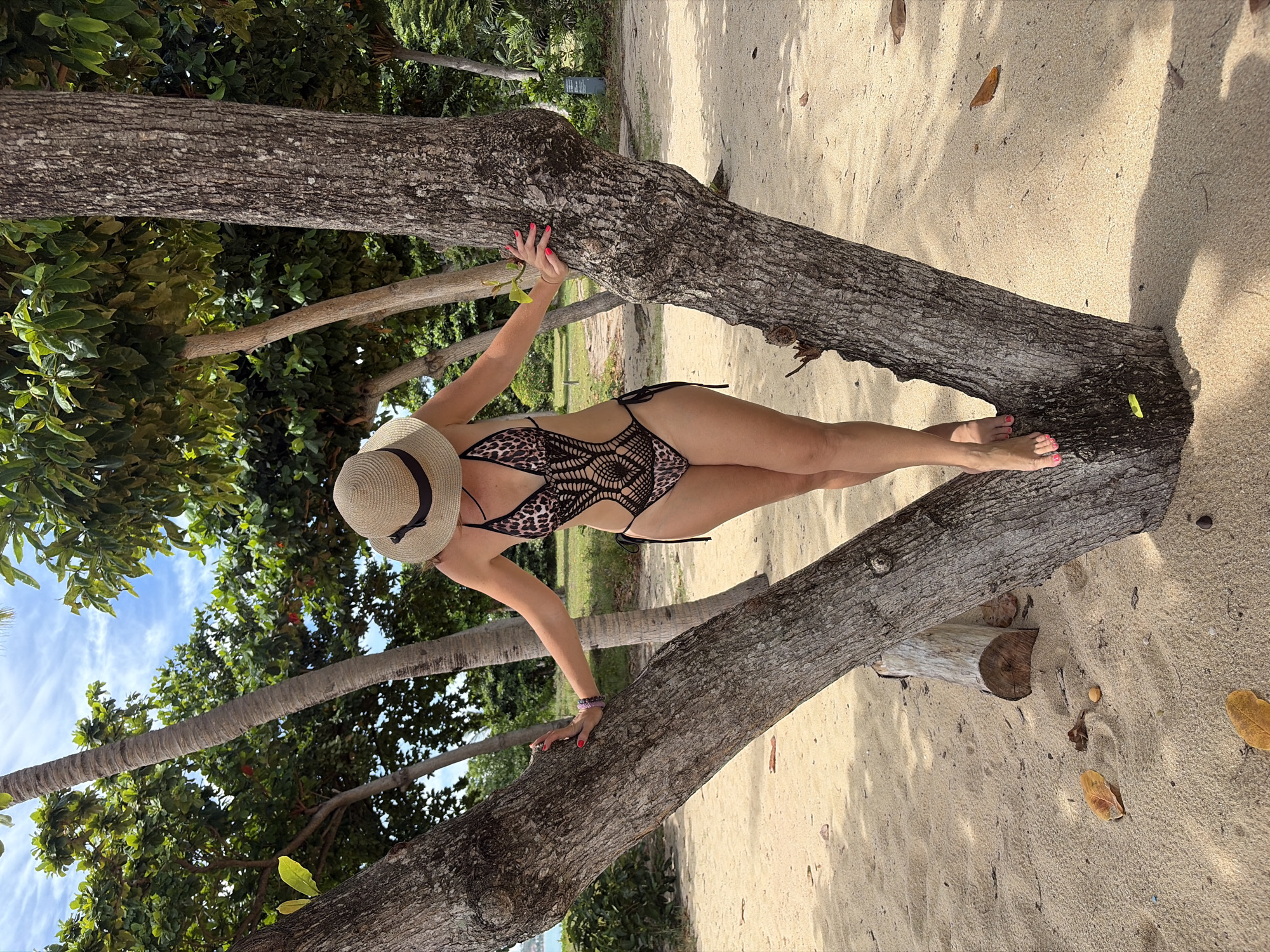 A woman wearing a wide-brimmed straw hat and a leopard-print one-piece swimsuit stands with her arms resting on a tree trunk. The swimsuit has cutouts and black crochet detailing. Her fingernails are painted bright pink. She is barefoot, with her feet positioned on a section of the tree. The scene is a sandy beach with lush green trees in the background, suggesting a tropical environment. The sky is partly cloudy. The sand has some fallen leaves scattered on it. The image appears to be taken in Baan Bo Phut, Thailand, a location known for its beaches and tropical flora.