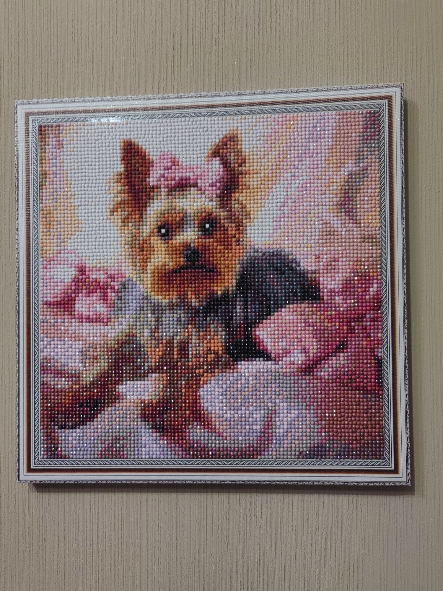 The image shows a framed diamond painting of a Yorkshire Terrier dog. The dog is depicted with a pink bow on its head and a dark collar. It is surrounded by flowers in shades of pink, white, and red. The diamond painting is hanging on a beige, textured wall, likely in a residential setting in the city of Ostafievo, Russia. The lighting suggests it is daytime, and the overall scene is static, with no visible people or actions taking place.