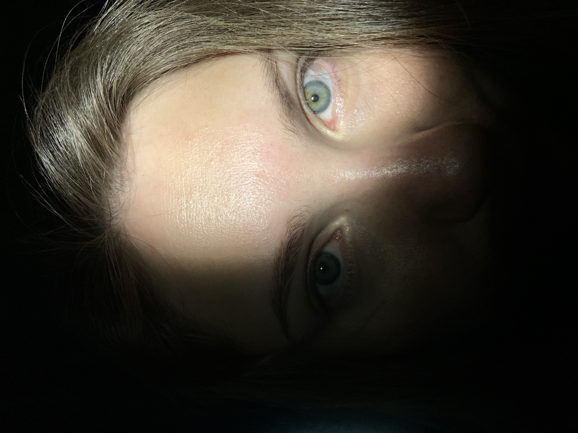 The image shows a close-up of a person's face, with an emphasis on the eyes. One eye appears to have a green hue while the other is blue. The lighting is dramatic, with a strong contrast between light and shadow, bisecting the face diagonally. Hair is visible at the top of the frame. The location is not discernible from the provided image. There is no information about the city of Chaikovsky, Russia in the image. There are no visible objects or discernible actions. The only interaction is the eye contact with the viewer.