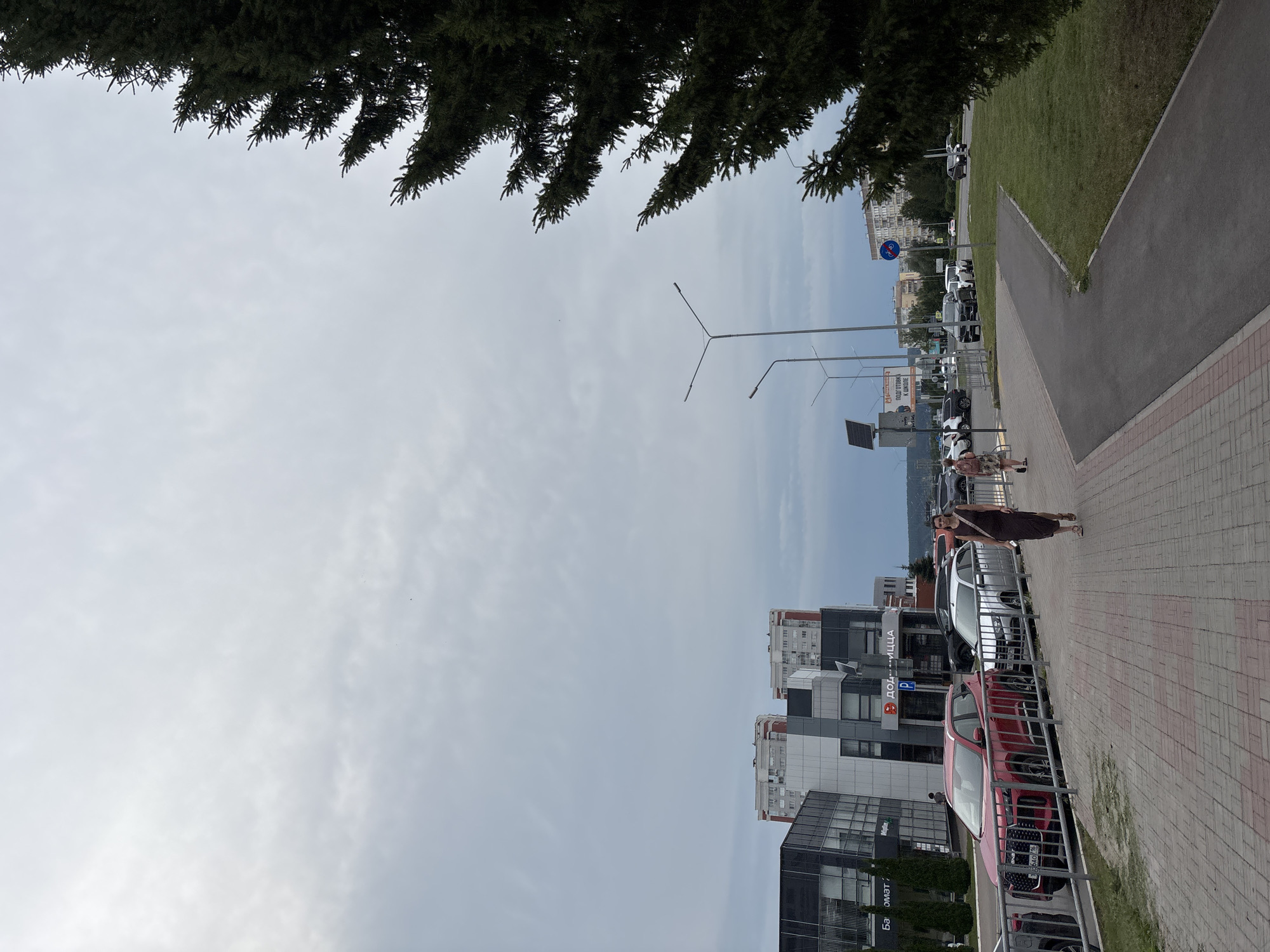 The image shows a sidewalk and street scene in Naberezhnye Chelny, Russia. On the right, a large evergreen tree dominates the foreground. A paved sidewalk runs parallel to a street where several cars are parked, including a red SUV and a silver sedan. Two women are walking on the sidewalk. In the background, there are modern buildings, some with glass facades, and residential apartment buildings. Streetlights are visible along the road. The sky is overcast.