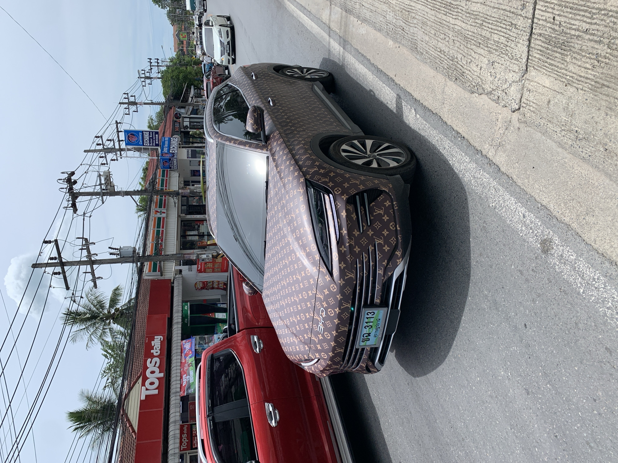 A brown Louis Vuitton patterned SUV is parked on the side of a street in Ban Bang Rak, Thailand. The SUV has a license plate with the number "3113" and Thai characters. A red pickup truck is parked to the left of the SUV. In the background, there are buildings with signs for "Tops daily" and "Exchange". Overhead power lines are visible across the sky. The street is paved with asphalt and there is a concrete sidewalk.
