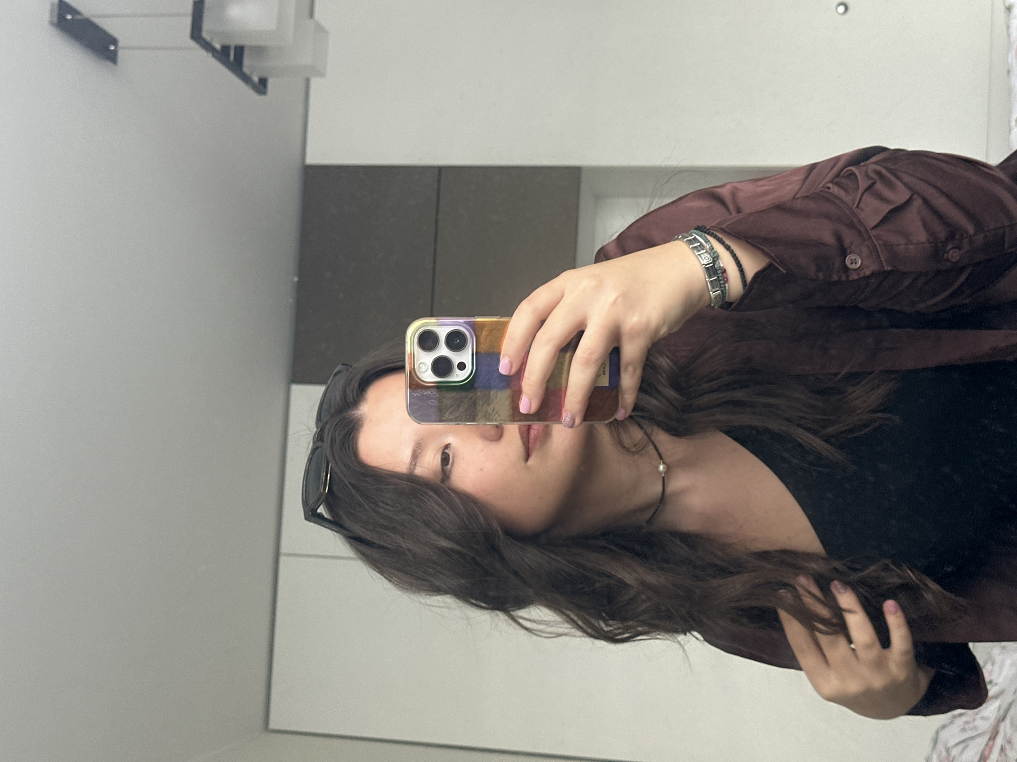A selfie captured in a mirror shows a woman holding a smartphone. She is facing the mirror with the phone covering part of her face. The woman has long, wavy brown hair, and she's wearing sunglasses pushed back on her head. She is also wearing a burgundy button-down shirt over a black top. On her left wrist, she has multiple bracelets. The background shows a wall, a light fixture, and a cabinet, suggesting an indoor location, possibly a bathroom or dressing room. There is no information about the city of Sofia, Bulgaria in the image.