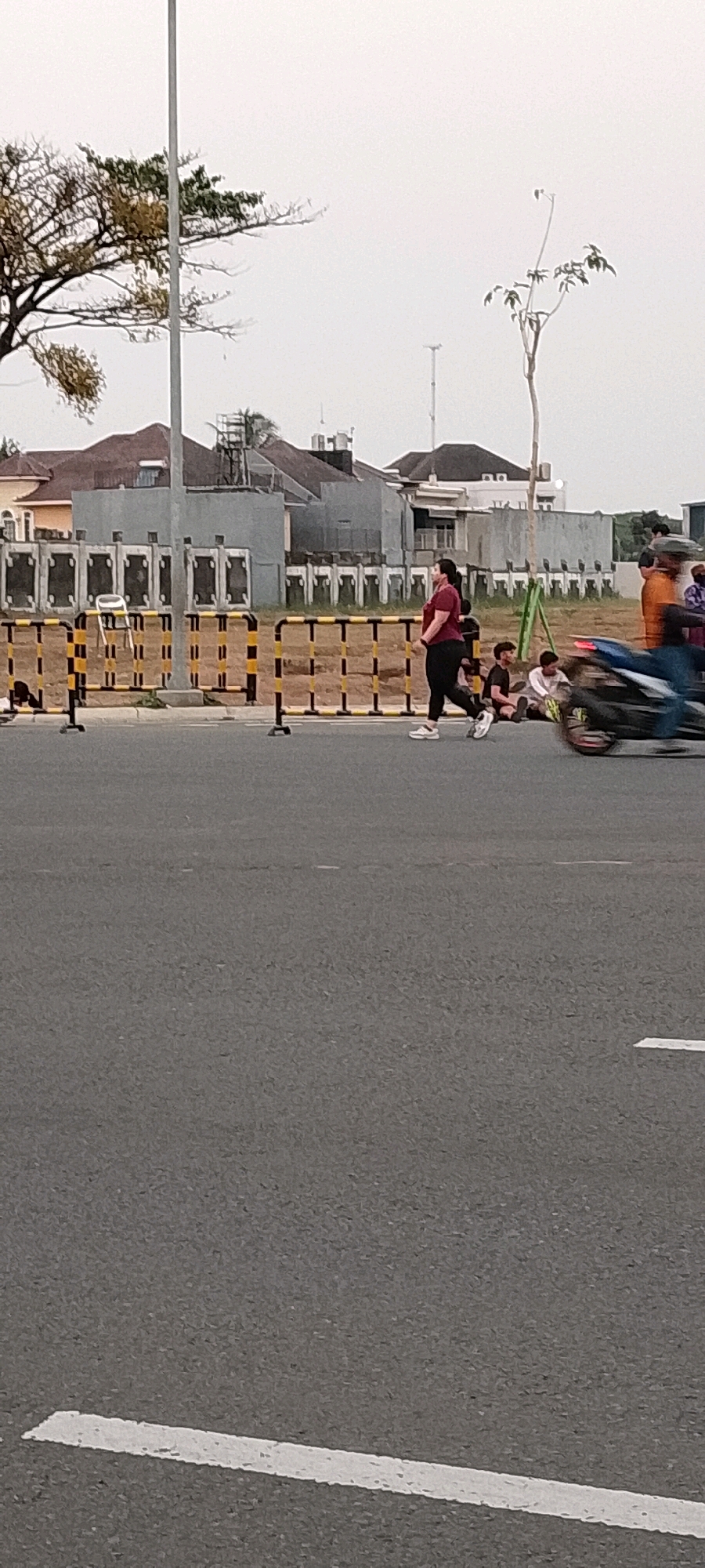 The image shows a street scene in Gunung Putri, Indonesia. In the foreground, there is a paved road with white lane markings. In the midground, a woman in a maroon shirt and black pants is walking across the street. She is wearing white sneakers. Behind her, several people are sitting on the ground. A motorcycle with a rider is passing by on the right side of the frame, appearing blurred due to motion. There is a yellow and black striped barrier in the background, separating the road from a construction area. Beyond the barrier, there are buildings and trees. The sky is overcast.