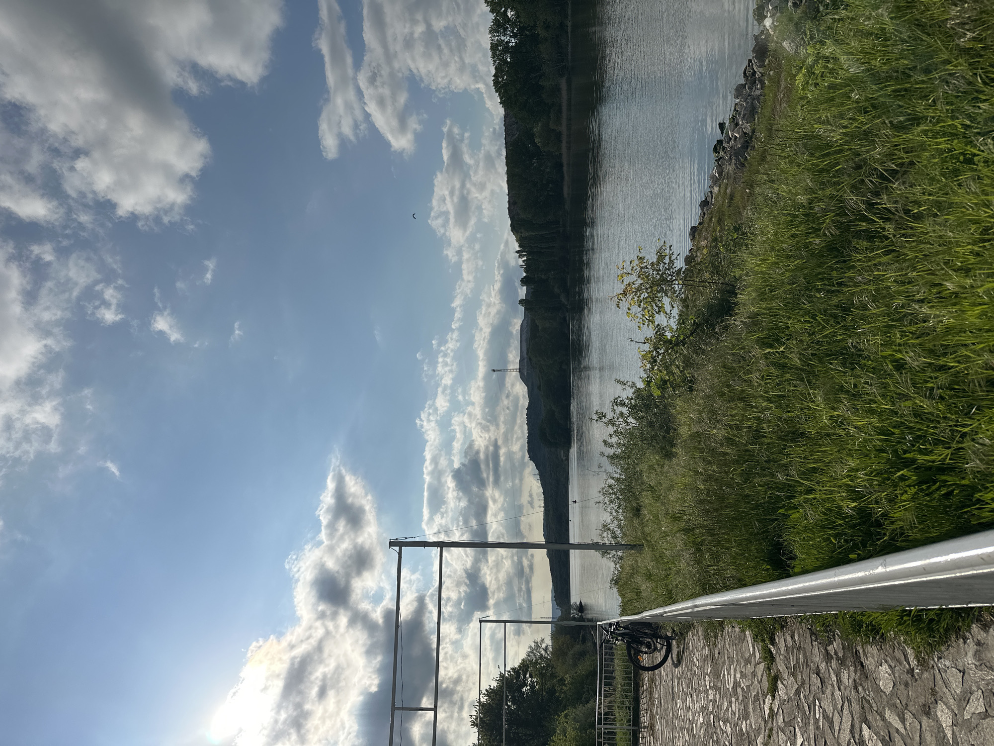 A landscape view captures a waterside location with a cloudy sky overhead. A body of water, possibly a lake or a river, occupies the right portion of the frame, its surface reflecting the sky. A verdant embankment runs along the water's edge, where tall green grass is visible. A paved path, made of stone, runs along the bottom. A guard rail stretches across the width of the photo and a bicycle is parked nearby. Further into the distance, a bridge tower can be seen on the other side of the water. In the sky are clouds and sunlight. The scene is likely in Beloslav, Bulgaria.