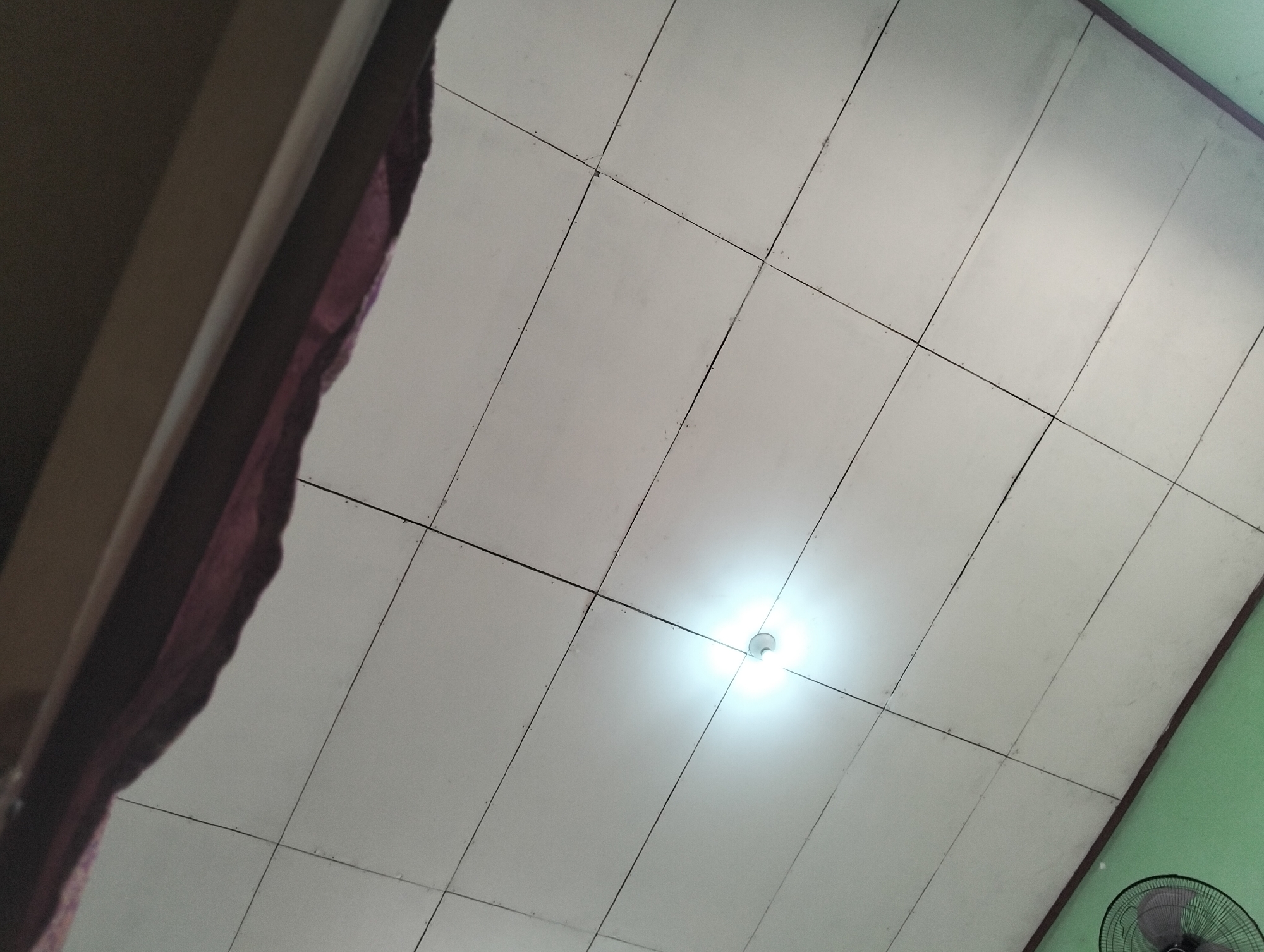 The image shows a ceiling with a light fixture and a portion of a wall. The ceiling is made of rectangular panels with visible seams. A bright lightbulb is visible in the center of the image, emitting a strong glare. To the left, a dark, draped fabric is partially visible, possibly curtains or a tapestry. On the right, a section of a green wall is present, with the top of a fan visible in the lower right corner. The scene appears to be indoors, likely in a room in Banjar Agung, Indonesia.