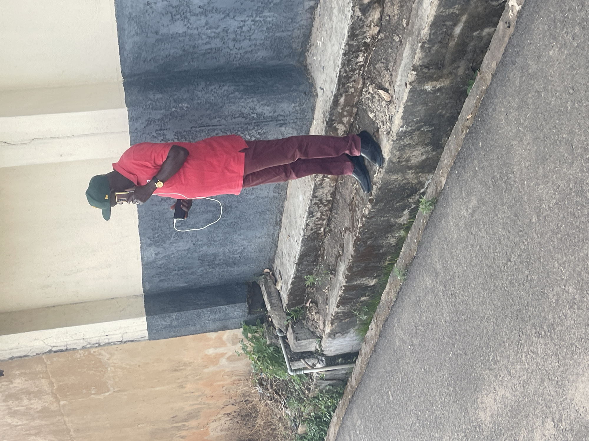 An adult man stands outdoors in a daylight scene, likely in an urban or semi-urban area of Jos, Nigeria. He is wearing a red polo shirt, maroon trousers, and black shoes, complemented by a dark green baseball cap and a gold-toned watch on his left wrist. The man is holding a dark-colored smartphone in his right hand, appearing to be either on a call or interacting with the device, as indicated by the white wired earphones plugged into it, with the cable dangling.
The man is positioned next to a multi-tiered concrete structure that shows signs of weathering, with small green plants growing from its cracks and crevices. To the viewer's right of the man is a paved surface, characteristic of an asphalt road or pathway. In the background, there is a cream-colored wall or building structure situated above a darker, possibly grey or black, lower wall or beam. The scene is brightly lit, indicating clear daytime conditions. No specific emotions are discernible on the man's face, and there is no visible text in the image. The setting suggests a functional or industrial part of a city, characterized by concrete infrastructure.