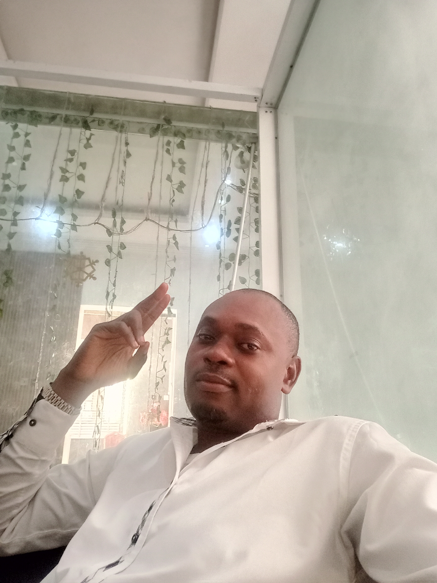 A medium shot captures a dark-skinned man indoors, likely in Gwarinpa, Nigeria. He is dressed in a white, collared button-up shirt with long sleeves, and a silver-toned watch adorns his left wrist. His right hand is raised in a gesture. The background reveals an interior space defined by white-framed glass or acrylic panels and a window or decorative element featuring strands of artificial ivy-like vines. A framed object is also visible through the transparent window treatment. The man is the focal point of the image.