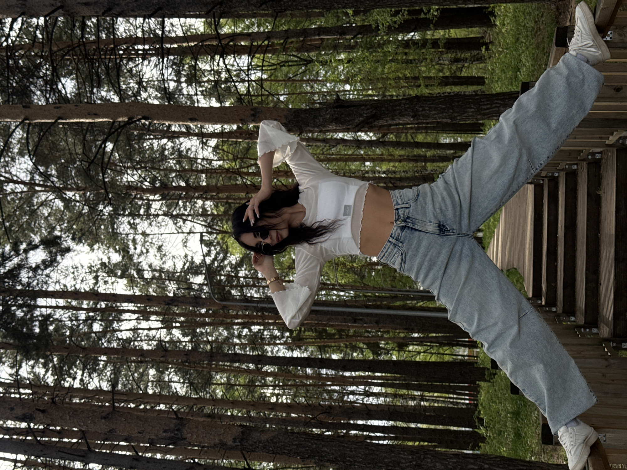 A woman poses in a forest in Chaikovsky, Russia. She wears a white cropped top, light blue baggy jeans, white sneakers, and sunglasses. She stands with one foot on a wooden stairway and the other extended upwards towards the trees. Trees surround her.