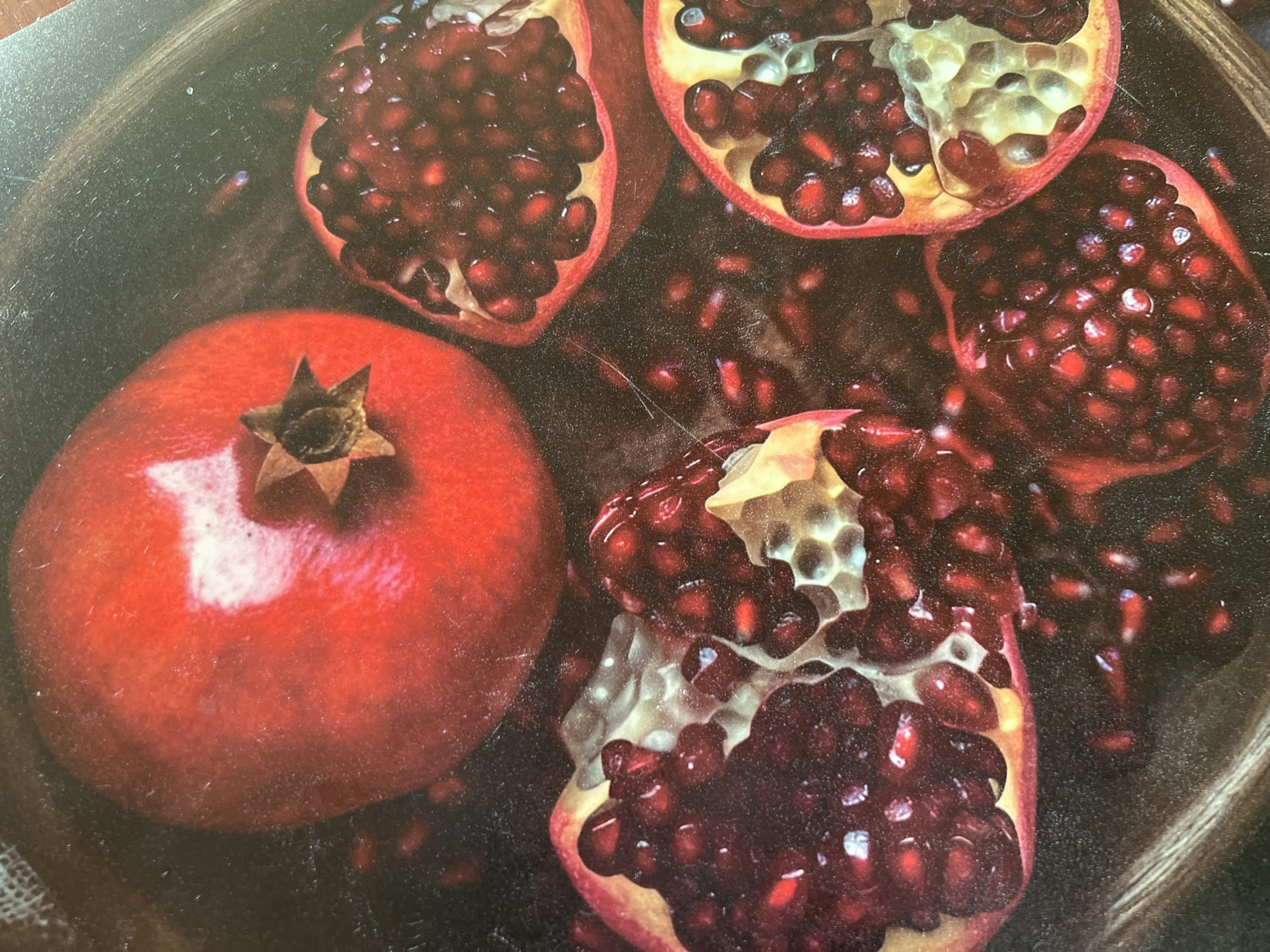 The image displays a close-up, overhead view of several pomegranates and pomegranate seeds scattered across a dark, textured surface, possibly a bowl or tray. One whole pomegranate, predominantly red with a star-shaped calyx, is visible in the upper left. To its right, a cut pomegranate reveals its jewel-like arils. Further to the right, another half-pomegranate shows its segmented interior filled with dark red seeds. In the lower portion of the image, two more open pomegranates are visible, one appearing to be a quartered section and another a larger half, both showcasing abundant red arils. The scene is lit to highlight the glossy texture of the fruit and seeds. There are no people or discernible city landmarks, and therefore no specific location within Minsk, Belarus can be identified.