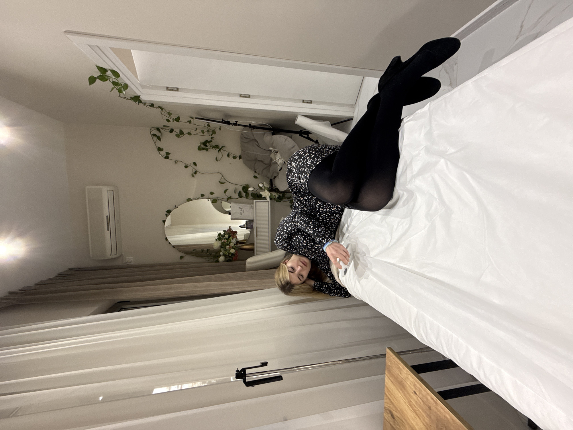 A blonde Caucasian female, appearing to be in her 20s to early 30s, is positioned on her left side on a white-covered surface, consistent with an examination or treatment bed. She is dressed in a black long-sleeved mini-dress with a white floral pattern and opaque black tights. Her head is supported by her left hand, and she holds a small white electronic device in her right hand. A light blue bracelet is visible on her left wrist.
The indoor scene is located in Budva, Montenegro. To the immediate left foreground, a section of a light-colored wooden table with dark metal legs is visible. Adjacent to the table, a tall, slender metal stand with a phone holder attachment is present. The background features a window covered by sheer white and beige blackout curtains. Centered in the background is a white vanity table with drawers, surmounted by a round mirror. A vase containing a mixed floral arrangement and a white product bag or box branded "KELUNE" are displayed on the vanity. An air conditioning unit is mounted high on the wall above the vanity. Green leafy vines are draped along the wall and around a white doorframe on the right. A gray upholstered chair and a collapsed or partially visible metal stand are situated near the door. The floor consists of white, marble-patterned tiles. A bright light source is visible near the ceiling.