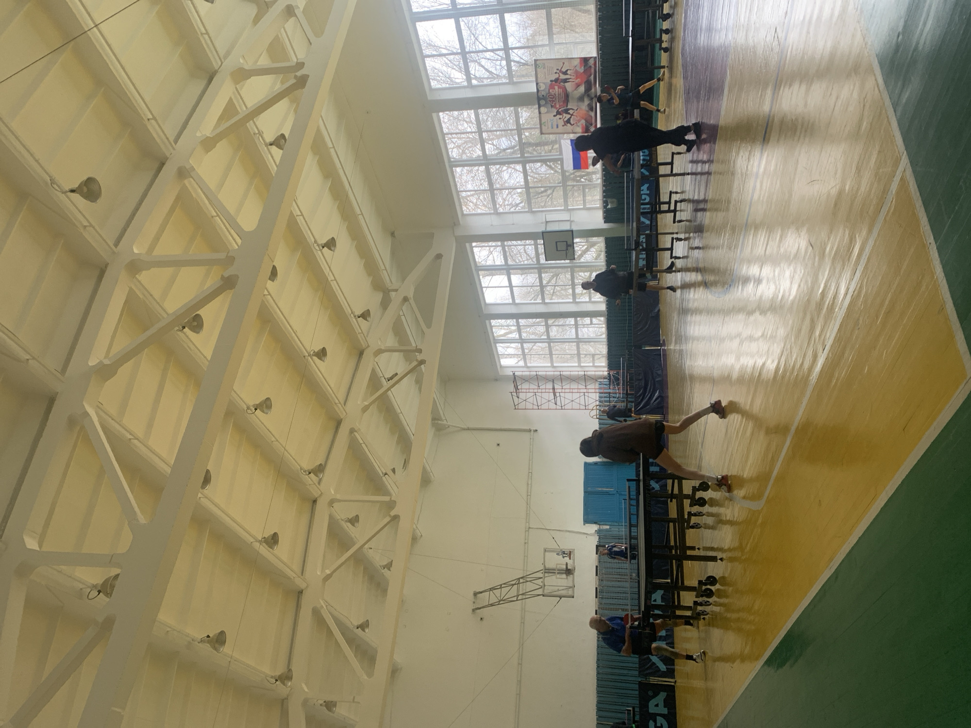 The image depicts the interior of a gymnasium, likely in Chaikovsky, Russia, based on the presence of the Russian flag. The gym features a high ceiling supported by white steel beams and is lit by rows of lights. Natural light enters through large windows at the far end. On the floor, several people are present. Some appear to be playing or practicing table tennis, while others are standing or walking. Table tennis tables are arranged in the gymnasium. The floor is marked with lines denoting different sections or areas, and a banner or poster is visible hanging on the wall near the windows.