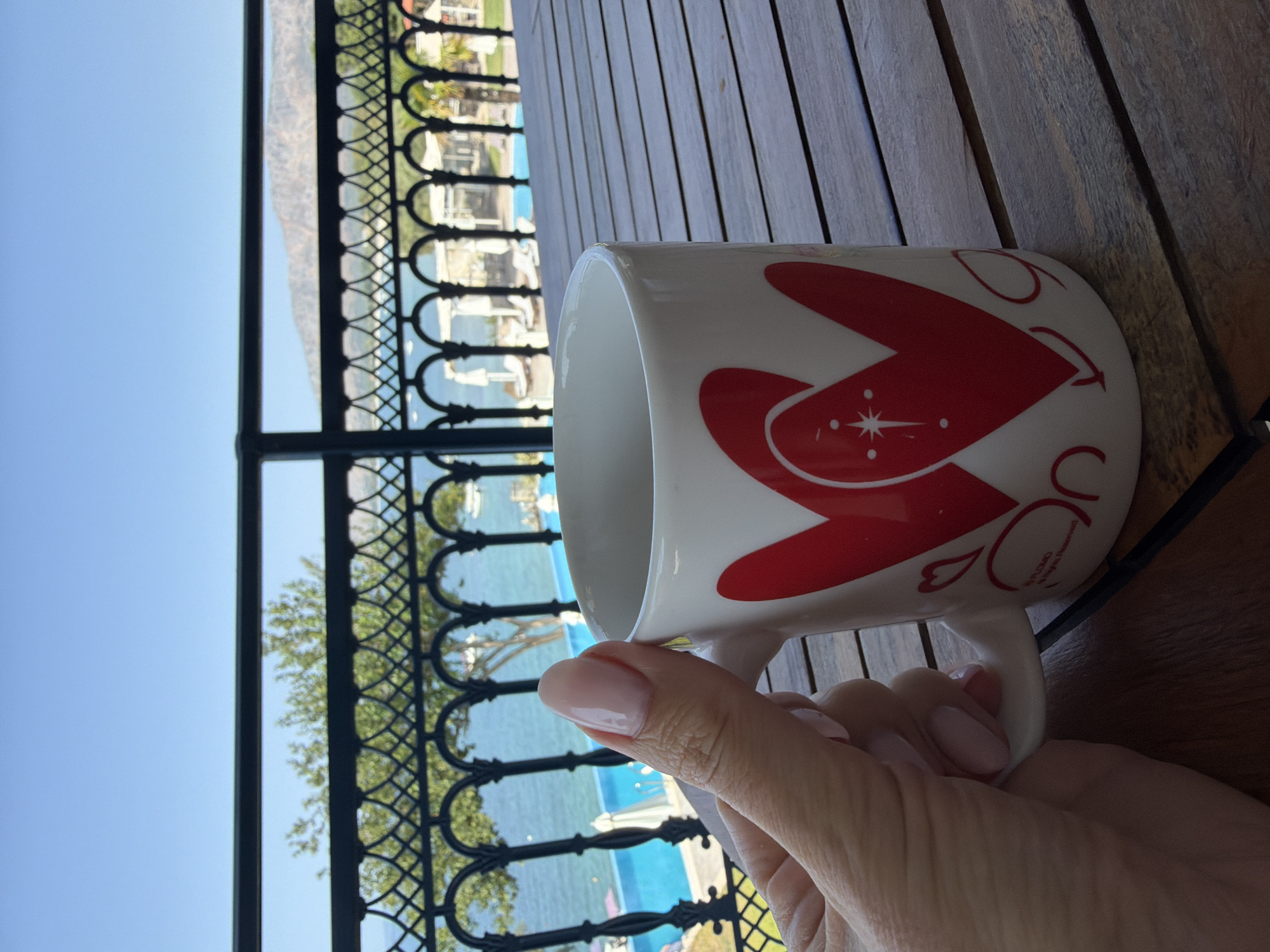A close-up, eye-level shot shows a person's hand holding a white mug with red heart designs, placed on a wooden table. The mug is partially in focus, with the hand and its clean, light pink-painted fingernails prominently displayed. In the background, a balcony railing with ornate black metalwork overlooks a bright blue swimming pool and a calm, blue sea. Beyond the pool and sea, a lush green tree is visible, followed by a distant mountain range under a clear blue sky. The scene suggests a serene outdoor setting, possibly a resort or a home with a view, located in the Municipal Unit of Alyzia, Greece, indicated by the presence of a pool and proximity to the sea and mountains.