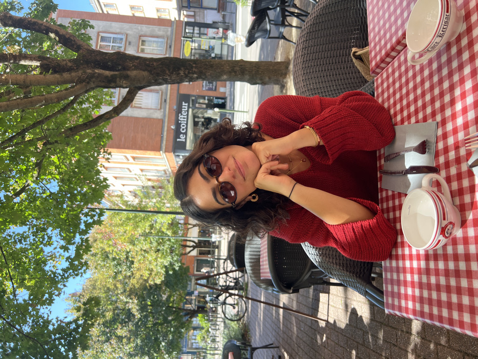 A young female with dark, wavy hair and oval-shaped brown sunglasses is seated at an outdoor café table, smiling towards the viewer. She is wearing a red knitted v-neck sweater, gold hoop earrings, a delicate gold necklace, and a gold-colored beaded bracelet on her right wrist, with a black elastic band on her left wrist. Her hands are clasped under her chin, resting on her forearms.
The table is covered with a red and white checkered tablecloth. On the table, to her left, is a white mug with red and black details, marked "2cl" and partially legible text "Orléans." To her right, a second white mug reads "Libre Kerisac." Between the mugs, a knife and fork are placed on a grey napkin. Dark rattan-style chairs with striped cushions are visible at her table and surrounding tables.
The scene takes place on a paved street or square under natural sunlight, with dappled shadows cast by overhead tree foliage. In the background, a multi-story building with reddish-brown brick upper floors and white window frames is prominent. The ground floor of this building houses a storefront with a sign that reads "Le Coiffeur" in white text on a dark background. A yellow sign is visible to the right of "Le Coiffeur." Further left, several bicycles are parked along the street. The location is **110 Rue de Bourgogne, Orléans, France**, identified by the specific architecture and "Le Coiffeur" sign, which corresponds to "Coiffeur le Coin" at this address.
