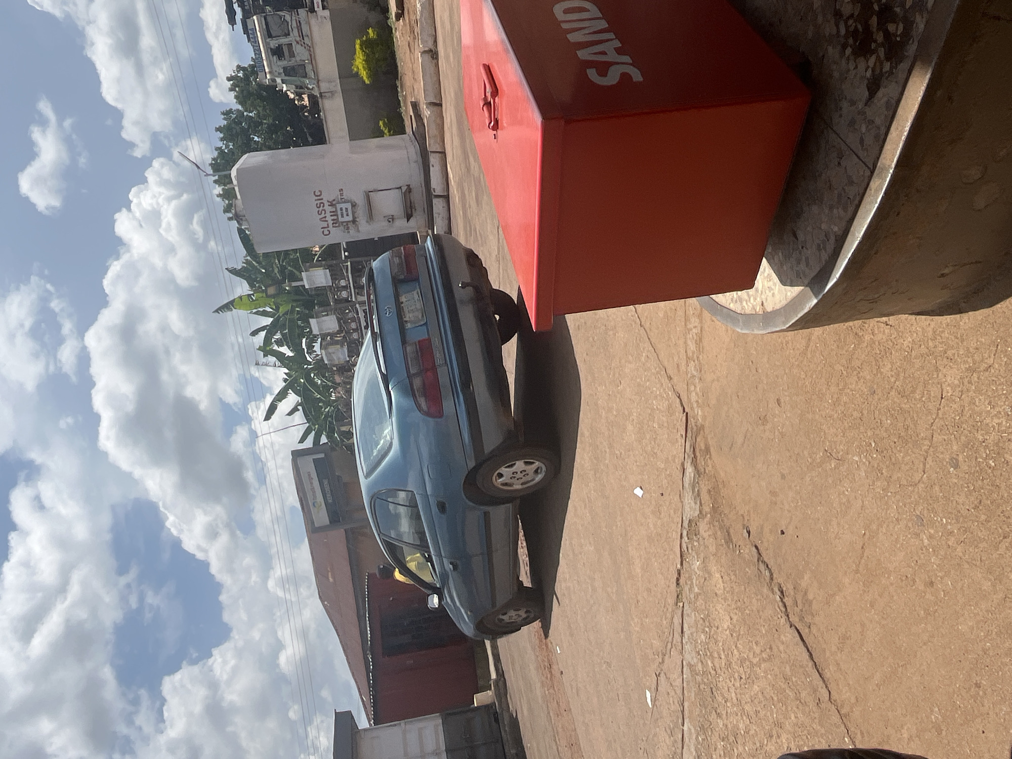 In the city of Gura Top, Nigeria, a teal sedan is parked on a dusty, cracked asphalt surface. The car is facing away from the viewer and is positioned to the left of a large, white, cylindrical tank labeled "CLASSIC BULK" in black lettering. To the right of the tank, a red, rectangular metal container with "SAND" printed in large white letters is visible. In the background, a red-roofed building with a sign that reads "TOTAL-NG KEROSENE" is present. Trees and other structures can be seen further in the distance. The sky is partly cloudy with white cumulus clouds against a blue background.