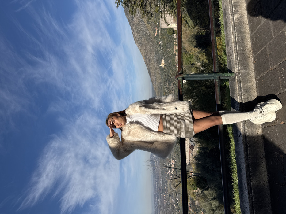 The image captures a young adult female standing on a paved scenic overlook in Tivoli, Italy, presenting a panoramic view of the Aniene valley. The individual, positioned slightly right of center, faces generally forward but looks towards the left, with her right hand raised to her forehead, shielding her eyes from direct sunlight. She is dressed in a cream-colored faux fur coat worn over a white ribbed mock-neck crop top, a brown and white plaid mini skirt, dark sheer tights, white knee-high socks, and chunky white athletic shoes. A dark metal railing supported by a concrete ledge horizontally spans the mid-ground, separating the foreground from the expansive valley. In the background, a clear blue sky with wispy white clouds dominates the upper frame. Below, the valley floor reveals a distant town or urban area on the left and a green, tree-covered hillside leading to a mountainous terrain on the right. A prominent yellow/ochre building, consistent with the Santuario di Ercole Vincitore, is visible in the middle distance of the valley towards the right. Scattered vegetation and other structures are discernable throughout the valley. Strong shadows are cast on the paved foreground, indicative of bright sunlight.