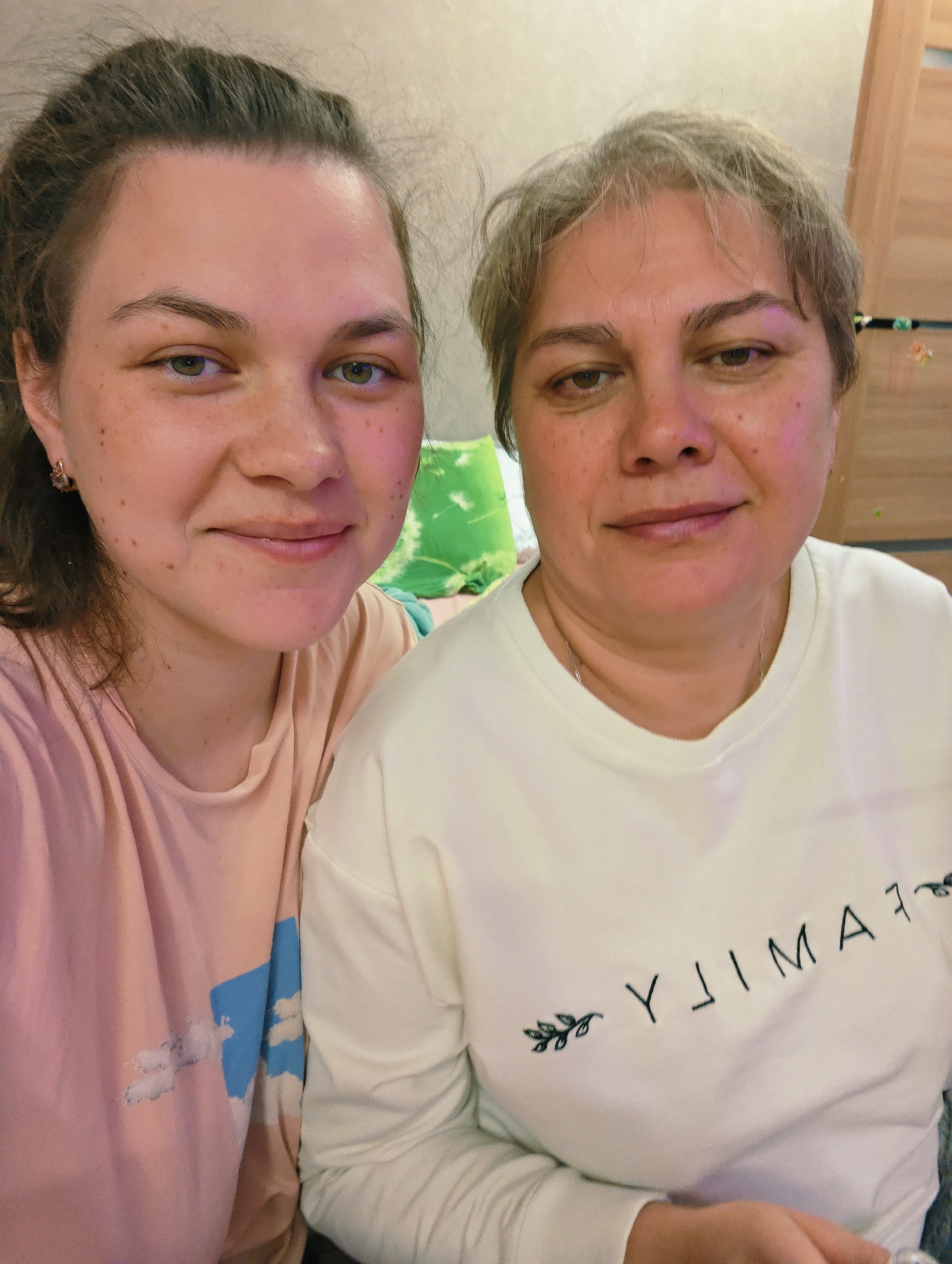 Two women are posing for a selfie indoors. The woman on the left has brown hair pulled back, green eyes, and freckles. She is wearing a pink t-shirt with a cloud design. The woman on the right has short, graying hair and brown eyes. She is wearing a white long-sleeved shirt that says "FAMILY" with floral embellishments. The word "FAMILY" is written backwards. The background includes a wall and what appears to be a wooden cabinet. Location is assumed to be in the city of Chaikovsky, Russia.