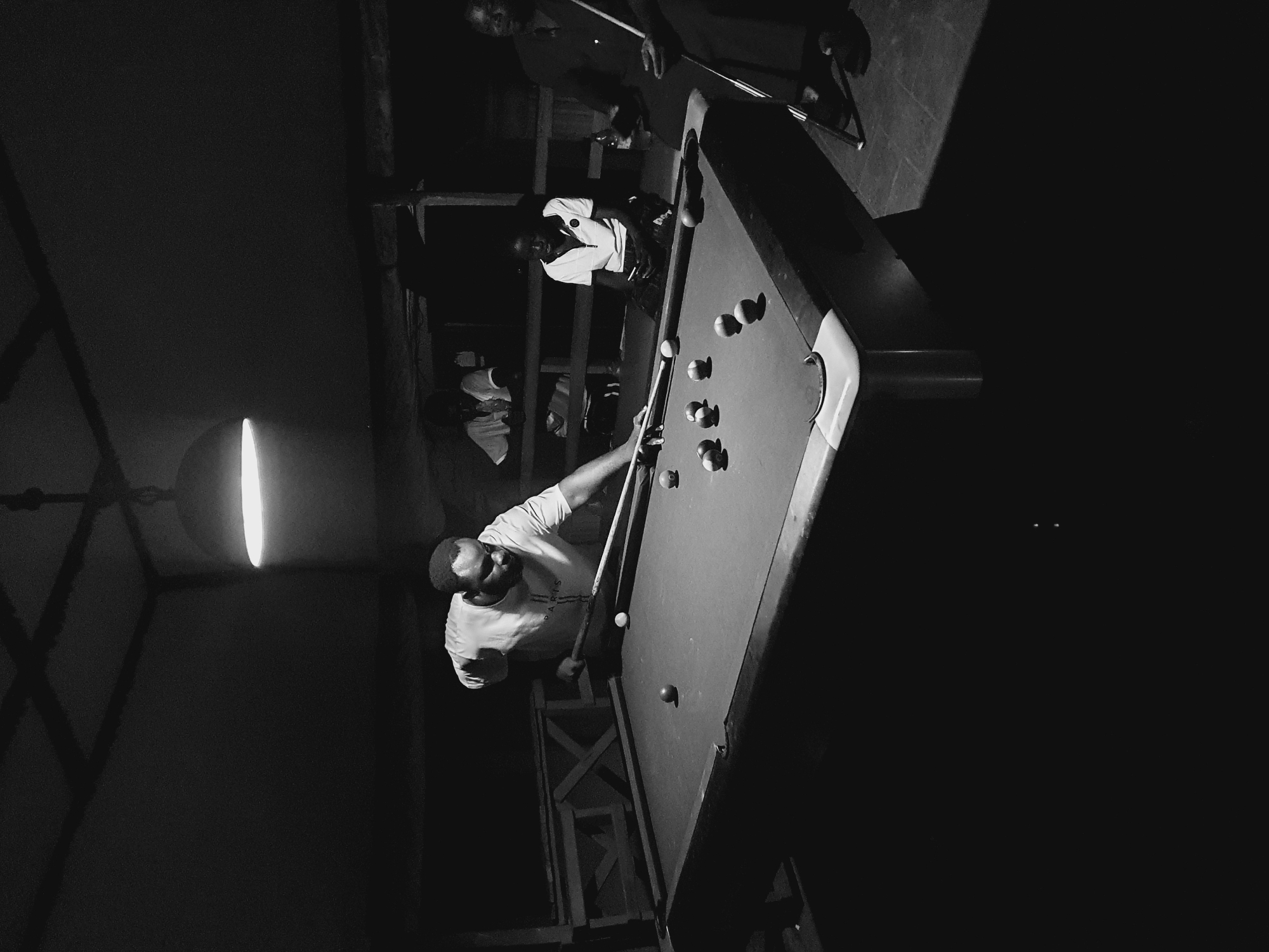 A black and white, low-light photograph captures a group of individuals engaged in a game of billiards in a dimly lit indoor space, possibly a bar or recreational area in Lokoja, Nigeria. A man in the foreground, wearing a t-shirt with the word "PARIS" visible, is actively playing, aiming his cue stick towards a cluster of billiard balls on the green felt surface. His posture suggests concentration as he is in the process of making a shot.
Several other people are present in the background, observing the game. One individual, a younger person in a white collared shirt, sits facing the pool table. Another person, dressed in a dark shirt and holding a cue stick, stands to the right, appearing to be waiting their turn or observing. Further back, another person is partially visible, leaning over a counter or railing.
The environment is characterized by wooden structures and a ceiling with visible beams, illuminated by a single overhead light fixture that casts a strong, focused glow on the playing area, creating dramatic shadows. The floor appears to be tiled or paved. The overall ambiance is one of casual recreation and social gathering in a somewhat rustic setting.