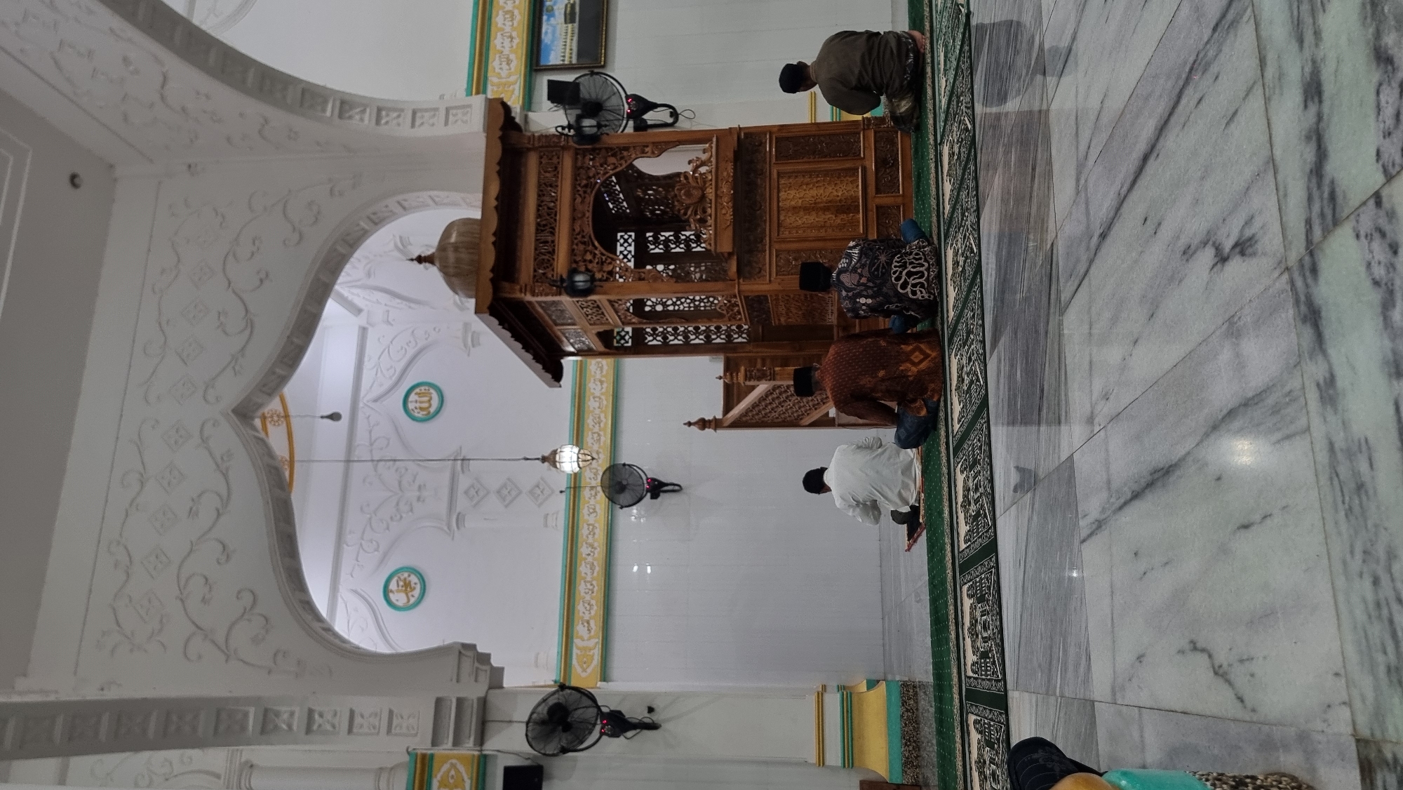 The image shows the interior of a mosque in Lapang Timu, Indonesia, during prayer. Four men are seated on green prayer mats on a patterned carpet, facing a wooden minbar. The men are dressed in traditional Islamic attire. The mosque features ornate white architecture with intricate carvings and decorative elements. Two black oscillating fans are mounted on the white walls. The floor is made of polished marble. A decorative wooden structure, likely the minbar, is positioned centrally. The lighting appears to be artificial, with a light hanging from the ceiling.