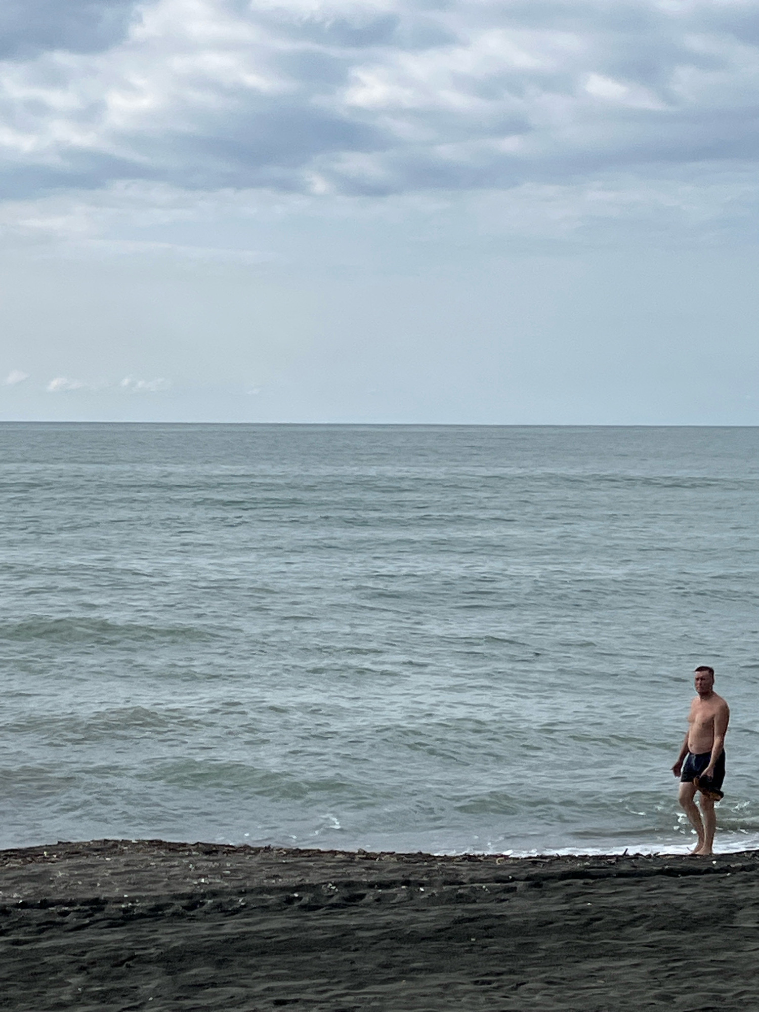 A man stands on the dark sand shore of the Black Sea in Ureki, Georgia. He is wearing dark swim shorts and holding a dark object in his right hand. The sea is calm with small waves gently lapping the shore. The sky above is overcast with a mix of grey and white clouds. The man is facing towards the left of the frame.