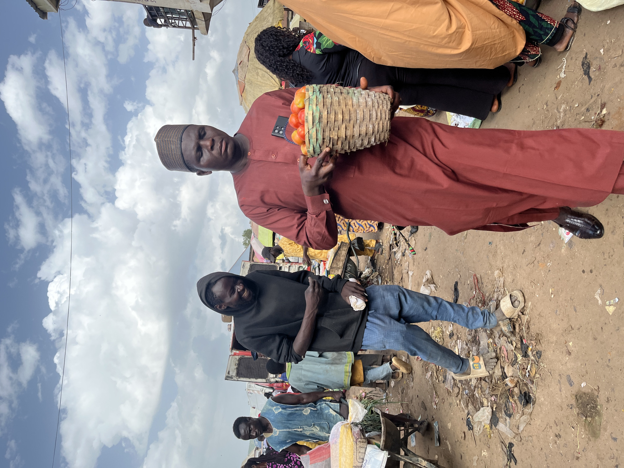 The image depicts an outdoor market scene in Rafin Par, Nigeria, under a partly cloudy sky. Two men are the primary subjects. The man in the foreground, wearing a dark hooded sweatshirt and blue checkered pants, is smiling and holding a small white object in his right hand. He is barefoot and standing amidst debris and trash on the ground.
Behind him and slightly to the left, another man is wearing a maroon traditional outfit and a patterned cap. He is holding a woven basket filled with tomatoes. His expression is neutral. The background reveals the bustling market environment with other individuals, stalls, and vehicles. The ground is dusty and littered with refuse, suggesting a typical market setting in a developing area. The lighting suggests it is daytime.