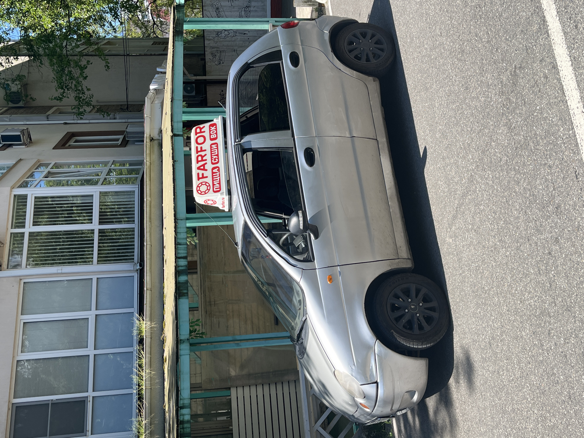 The image captures an outdoor scene on a sunny day in what appears to be an urban or suburban setting, specifically Sochi, Russia. A silver five-door hatchback car, likely a delivery vehicle, is prominently featured, parked or moving on an asphalt road.
The car is silver and has black multi-spoke rims. A red and white illuminated sign is mounted on its roof. The sign clearly displays the text "FARFOR" in large, stylized red letters with a gear-like graphic replacing one of the 'O's. Below "FARFOR," the Cyrillic text "ПИЦЦА СУШИ ВОК" is visible, which translates to "PIZZA SUSHI WOK," indicating the type of food offered by the establishment. A smaller web address, "farfor.ru," is also present at the bottom of the sign.
To the left of the car, a multi-story building stands with a mix of architectural styles. It features numerous windows, some with white frames and internal blinds, and others with horizontal panes. Several external air conditioning units are mounted on the building's facade. A distinctive feature is a light green-painted metal framework or balcony structure running along the building's side, partially covered by foliage and small plants growing in cracks. Above this structure, a textured gray wall or frieze is visible, adorned with what appear to be cartoonish food-related drawings. Green tree branches with dense foliage extend into the frame from the top left, casting shadows.
The ground consists of a textured asphalt road, with a white painted line visible on the right side of the image. Strong shadows cast by the car and other elements suggest bright sunlight, indicative of midday or early afternoon conditions with clear skies. No people are visible in the photograph, creating a calm and static atmosphere.