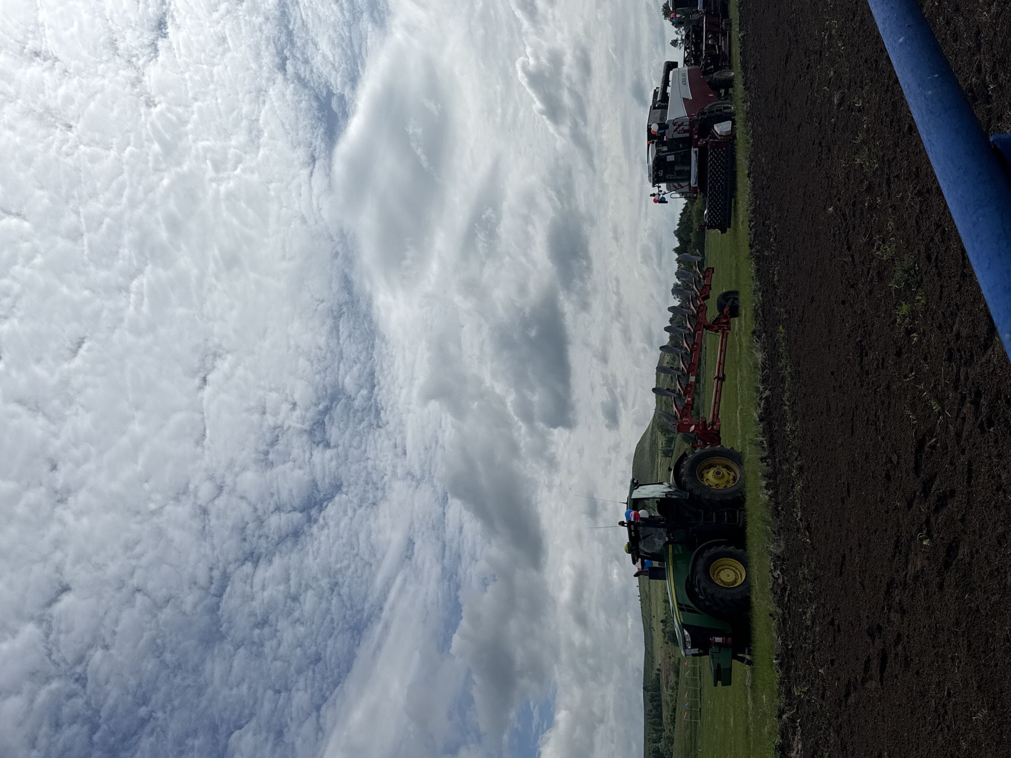 The image depicts a farm field under a cloudy sky. In the foreground, freshly tilled dark brown earth is visible. Further back, a green John Deere tractor with yellow wheels is parked on a grassy field, connected to a red plow. To the right of the tractor, a large red and white combine harvester is also positioned on the grass. The background shows rolling green hills with scattered trees. The scene is set in the city of Nurkeevo, Russia. There are no people clearly visible in the image.