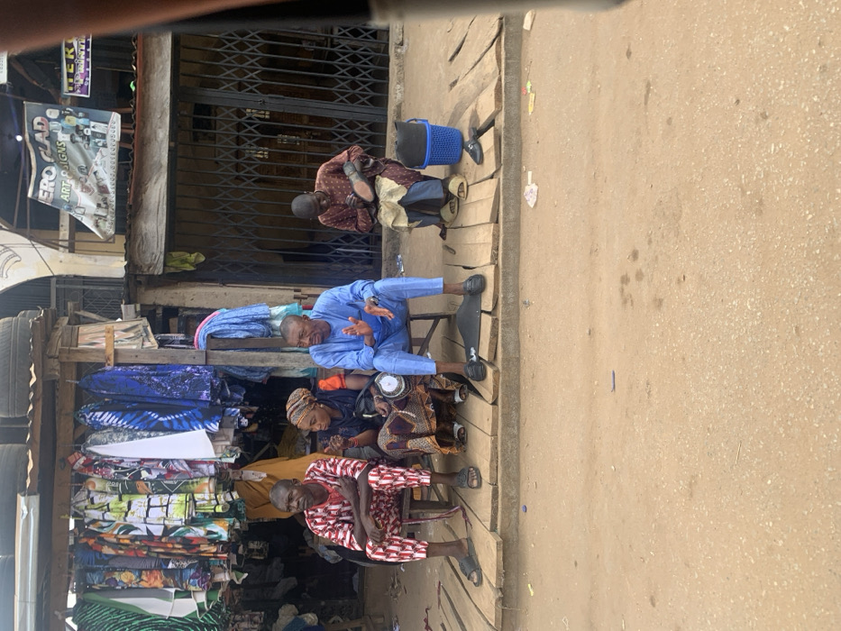 This image depicts a street scene in Egbedi, Nigeria, focusing on a raised wooden platform along a commercial thoroughfare. The foreground consists of a dirt road with scattered small debris.
On the wooden platform, four individuals are seated on low stools. From left to right:
1. A male, dressed in a red and white patterned traditional tunic and trousers, sits with arms crossed, observing. He wears black sandals.
2. A female, wearing a headscarf, a brown and orange patterned dress, and an orange long-sleeved top, gestures with her right hand while holding a small white disc-like object in her left, appearing to communicate. She wears white sandals.
3. A male, dressed in a light blue traditional tunic and trousers, sits with hands slightly open, looking forward. He wears black loafers.
4. A male, wearing a dark red patterned traditional tunic and trousers, sits with a blue cloth draped over his lap, actively shining a black shoe held in his hands. He wears light brown slides. A blue plastic basket containing a dark object is beside him, and another black shoe rests on the platform. A black rectangular mat or kit is positioned on the platform in front of the blue-clad man.
In the background, to the left, a retail stall displays numerous patterned garments hanging for sale beneath a corrugated metal roof topped with several automobile tires. To the right, a shop entrance is secured by a closed metal accordion gate. Above this shop, a banner reads "HERO GLAD ART SIGNS."