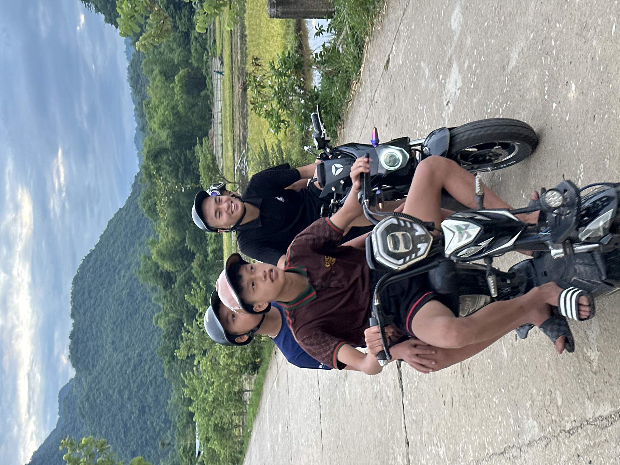 In the city of Chiềng Châu, Vietnam, three young males are riding on two electric scooters on a paved road. The lead scooter is ridden by a young male wearing a brown polo shirt with green and red trim, black shorts with red stripes, and black and white striped slides. He has a brown baseball cap on backwards. He is looking forward. Behind him, on the same scooter, is another young male wearing a dark blue t-shirt and a dark grey helmet. He is looking forward and slightly to his right. On the second scooter, the rider is a young male wearing a black polo shirt and a dark grey helmet. He is smiling and looking forward. The background of the image shows a rural landscape with lush green mountains, dense trees, and a rice paddy. The sky is overcast with some patches of light.